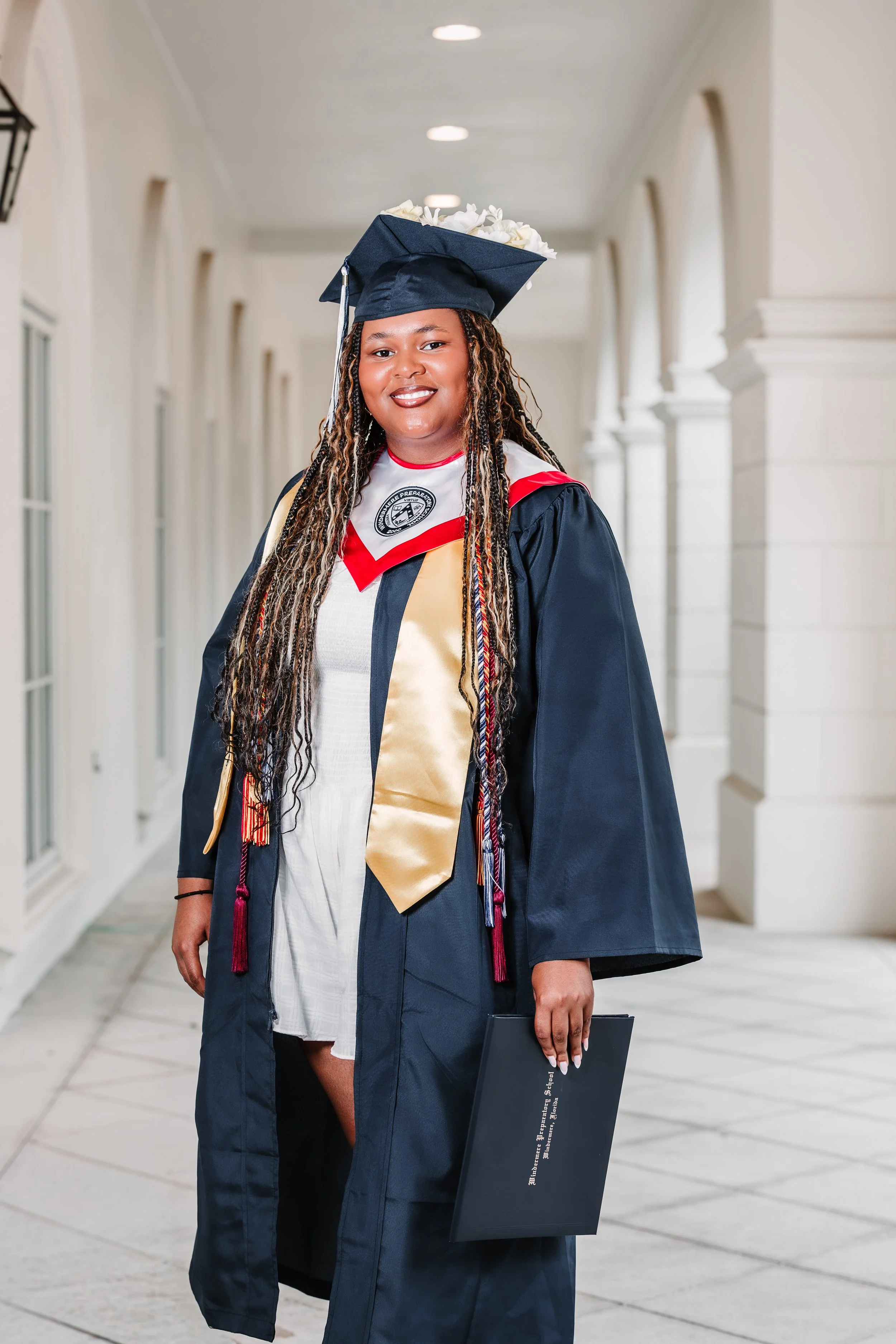 A woman in graduation cap and gown holding a diploma standing in a corridor.
