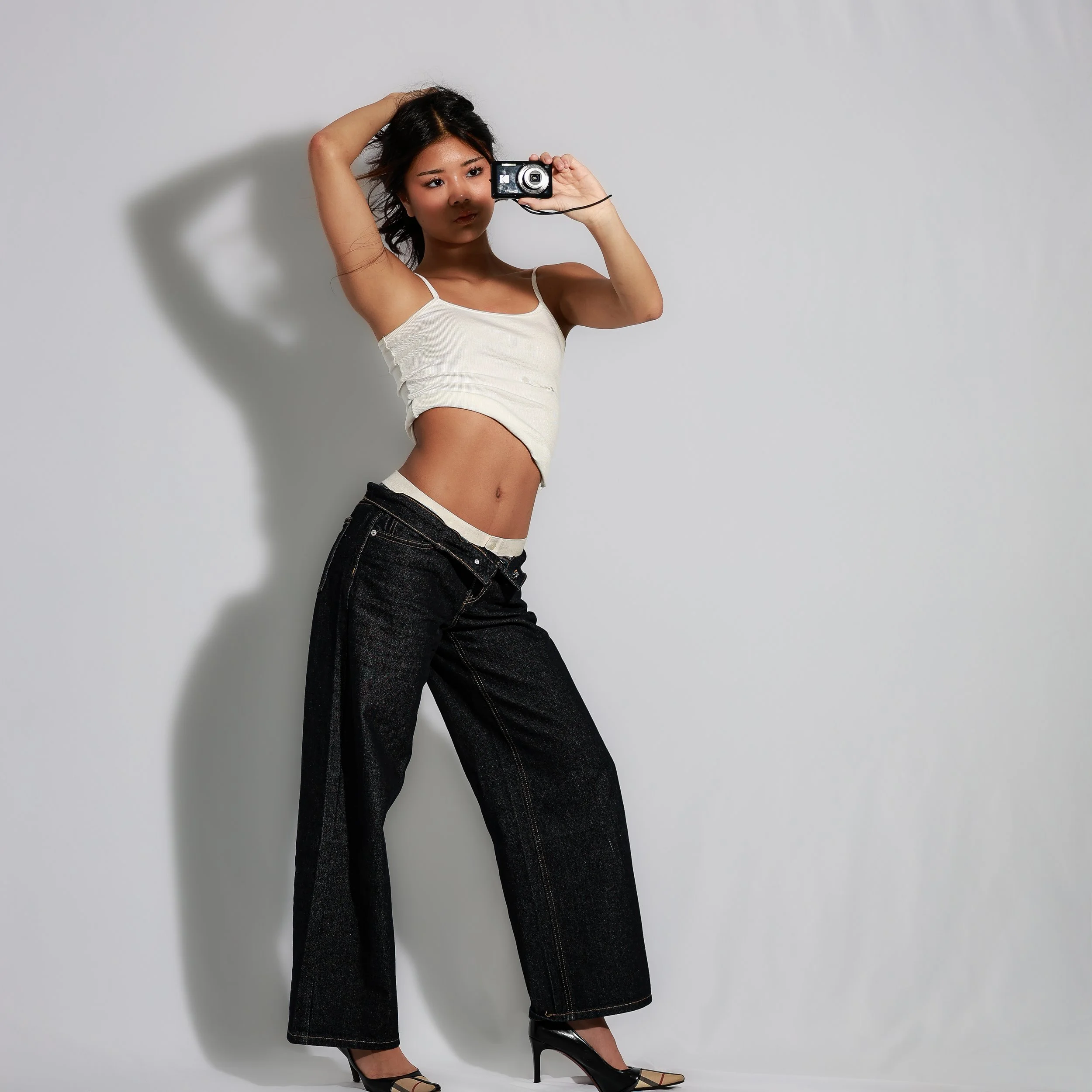 A woman taking a mirror selfie with a camera in her right hand on a plain light grey background. She is wearing a white crop top, black jeans, and black high heels. Her left hand is on her head, and her hair is loose.