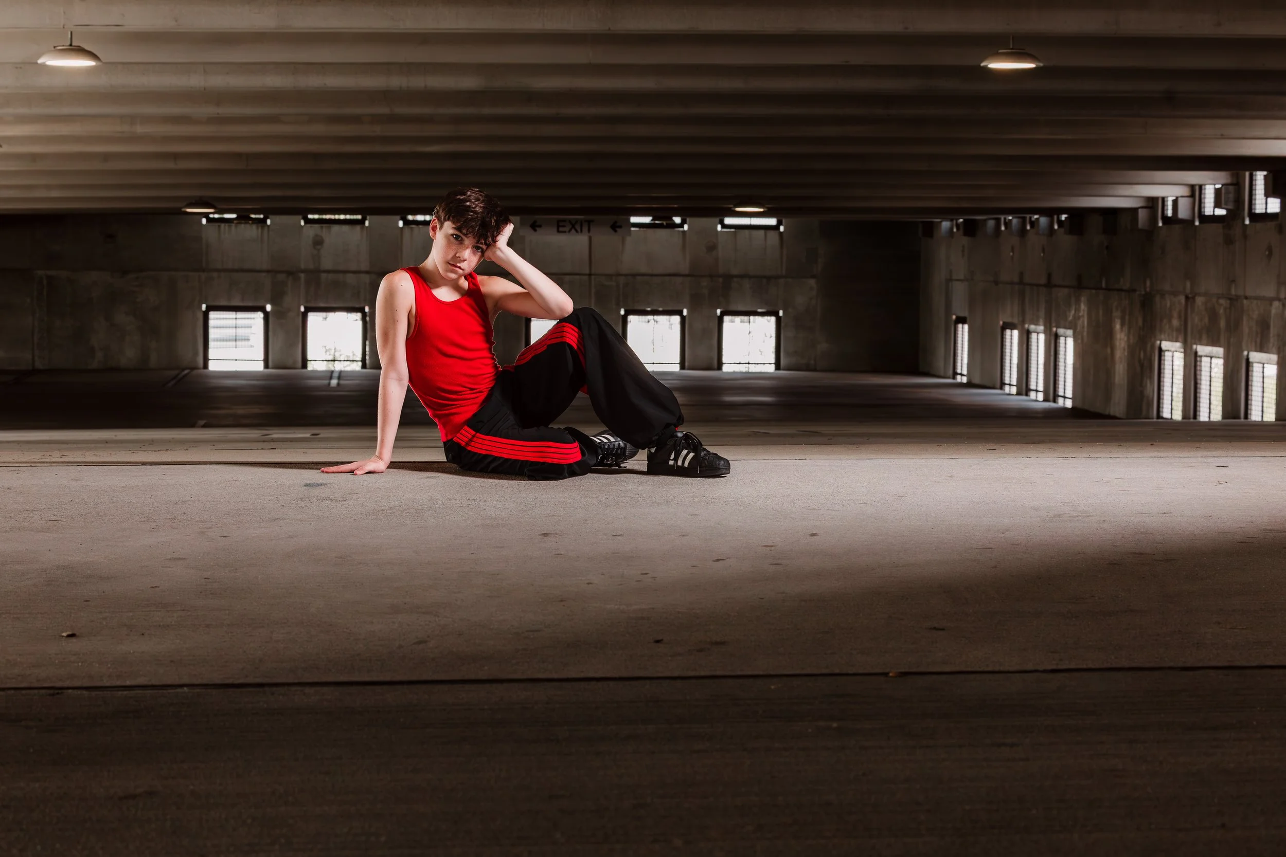 A young person in a red tank top and black sweatpants with red stripes, sitting on the ground in an empty parking garage, looking at the camera with a serious expression.