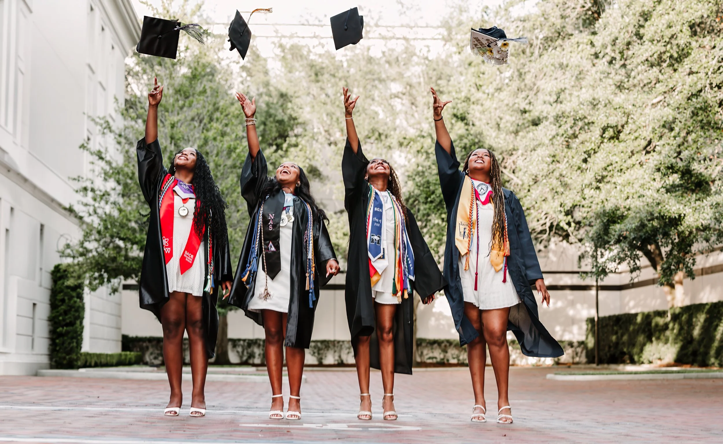 Four women in graduation gowns and caps are celebrating outdoors, tossing their caps into the air with a background of trees and a white building.