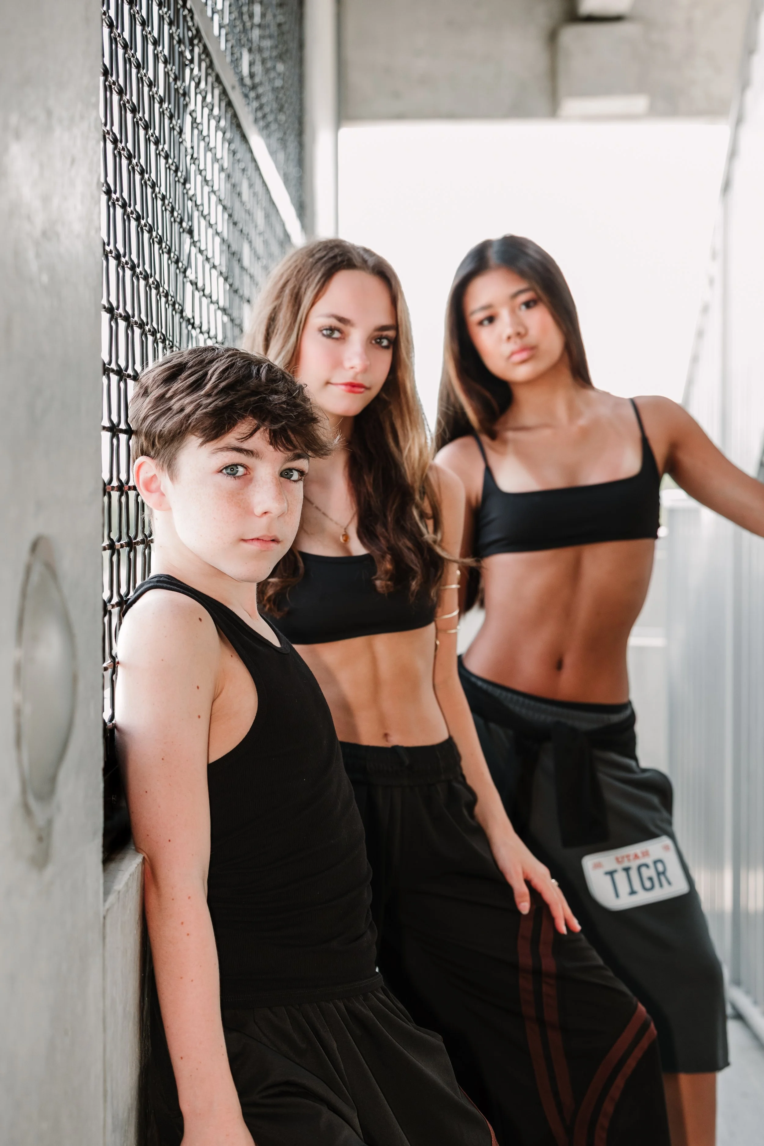 Three young women in black athletic wear standing against a concrete wall with a chain-link fence, leaning and looking at the camera, with a Utah license plate visible in the background.