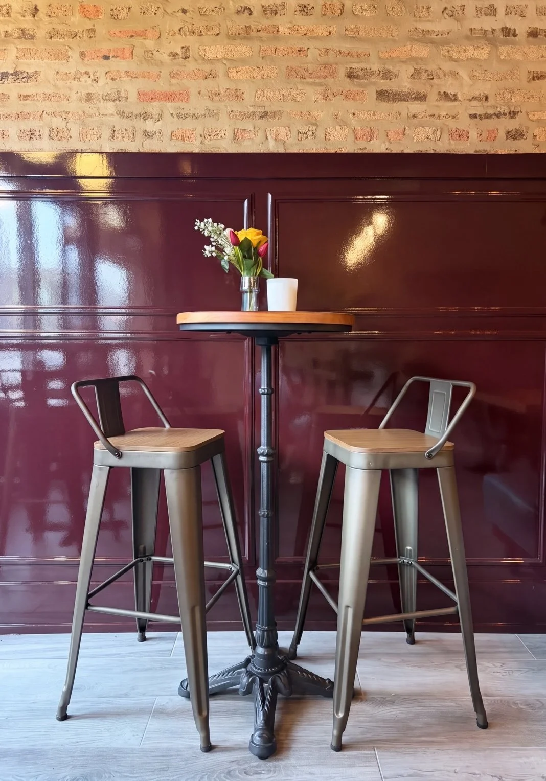 A small round table with a vase of yellow and pink tulips and a white candle, positioned between two metal bar stools with wooden seats, in a cozy cafe or restaurant with a brick wall and wooden paneling.
