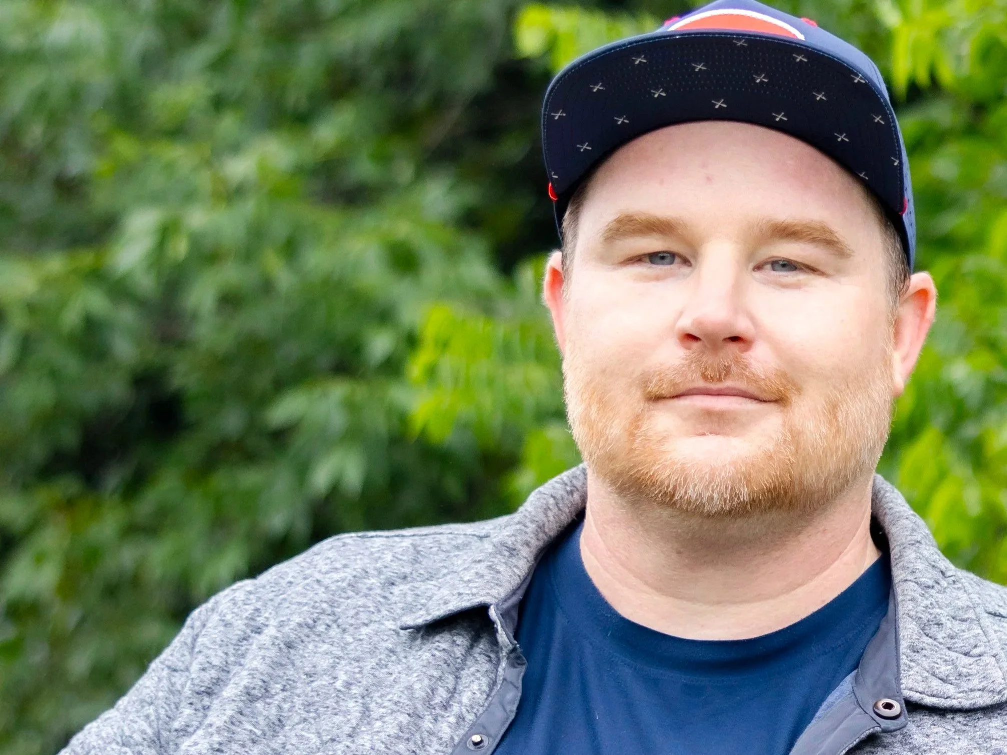Close-up photo of a young man with light skin, blue eyes, and a light beard, wearing a navy blue cap with small patterns, standing outdoors with a blurred green leafy background.