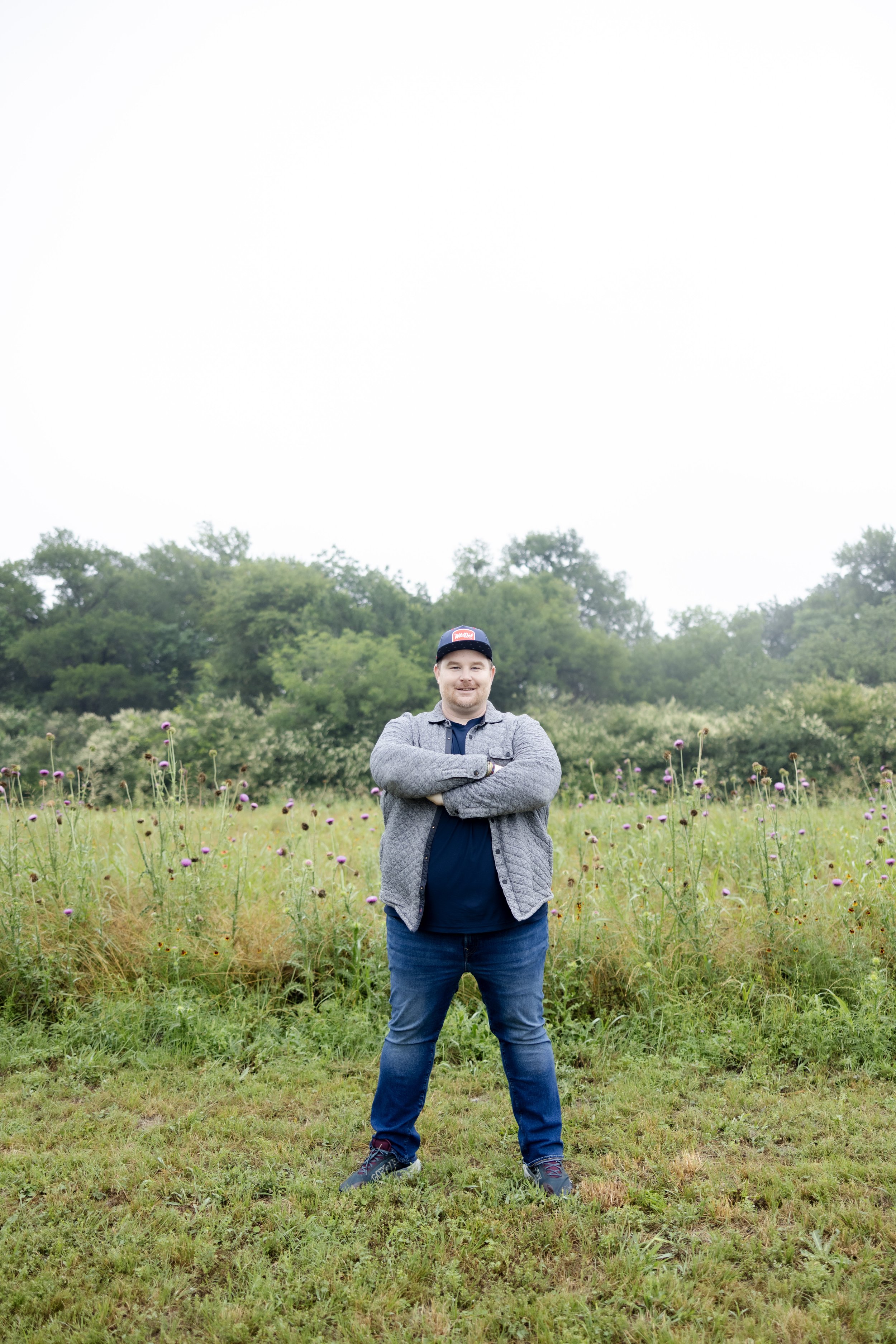 A man stands in a grassy field with purple flowers, arms crossed, wearing a gray jacket, blue jeans, and a baseball cap, with a backdrop of green trees under a cloudy sky.