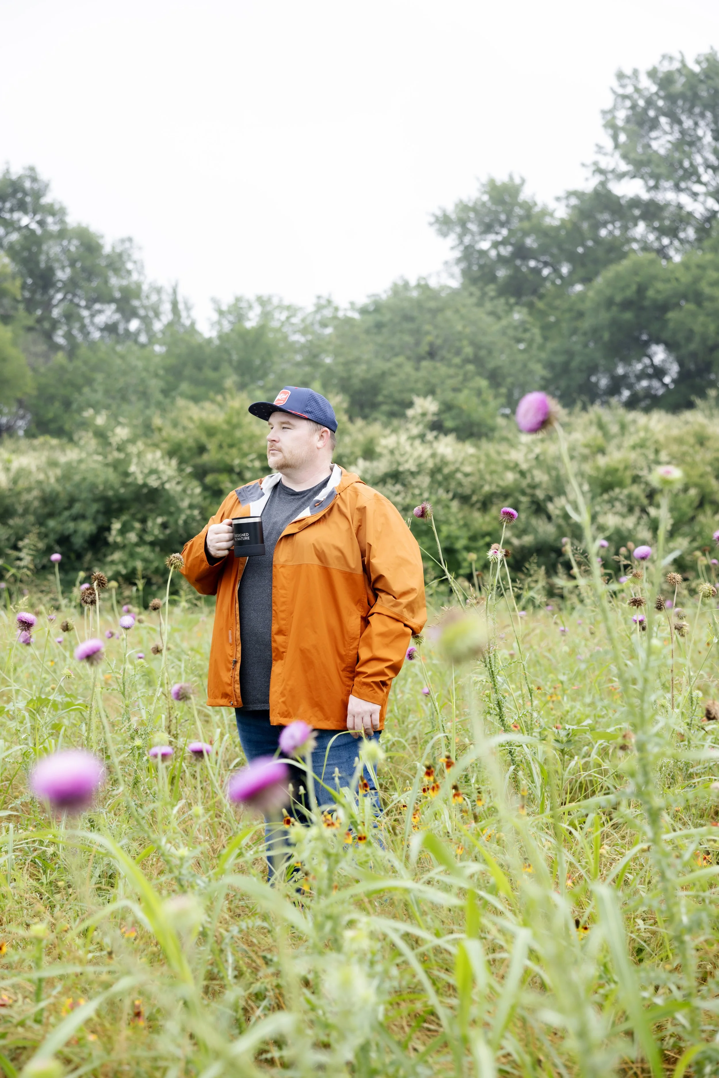 A man with a red beard and blue cap stands in a field of purple flowers, holding a black mug, wearing an orange jacket and looking to his left with a neutral expression. The background features green trees and an overcast sky.