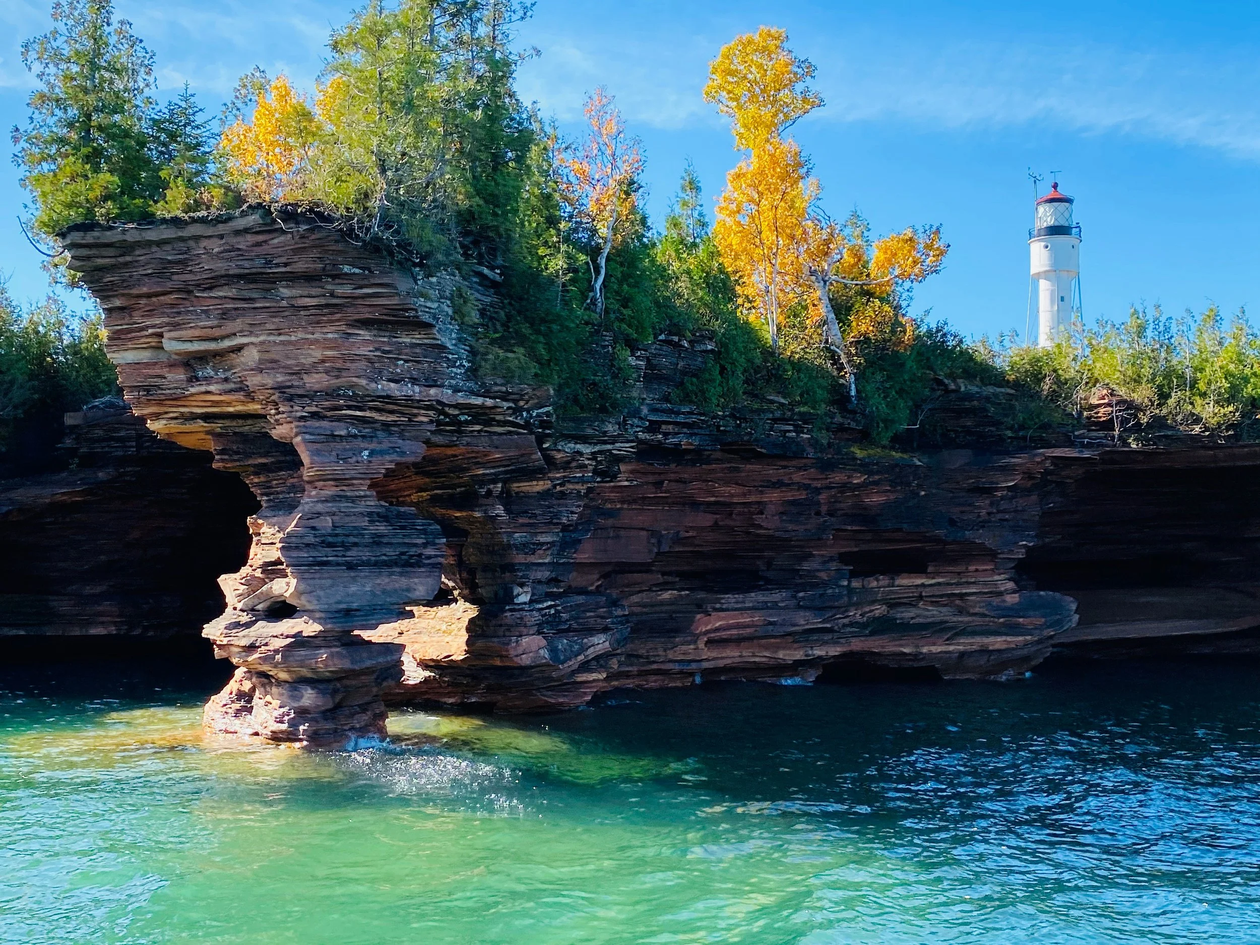 Rock formation with trees on top next to a lighthouse by the water under a blue sky.