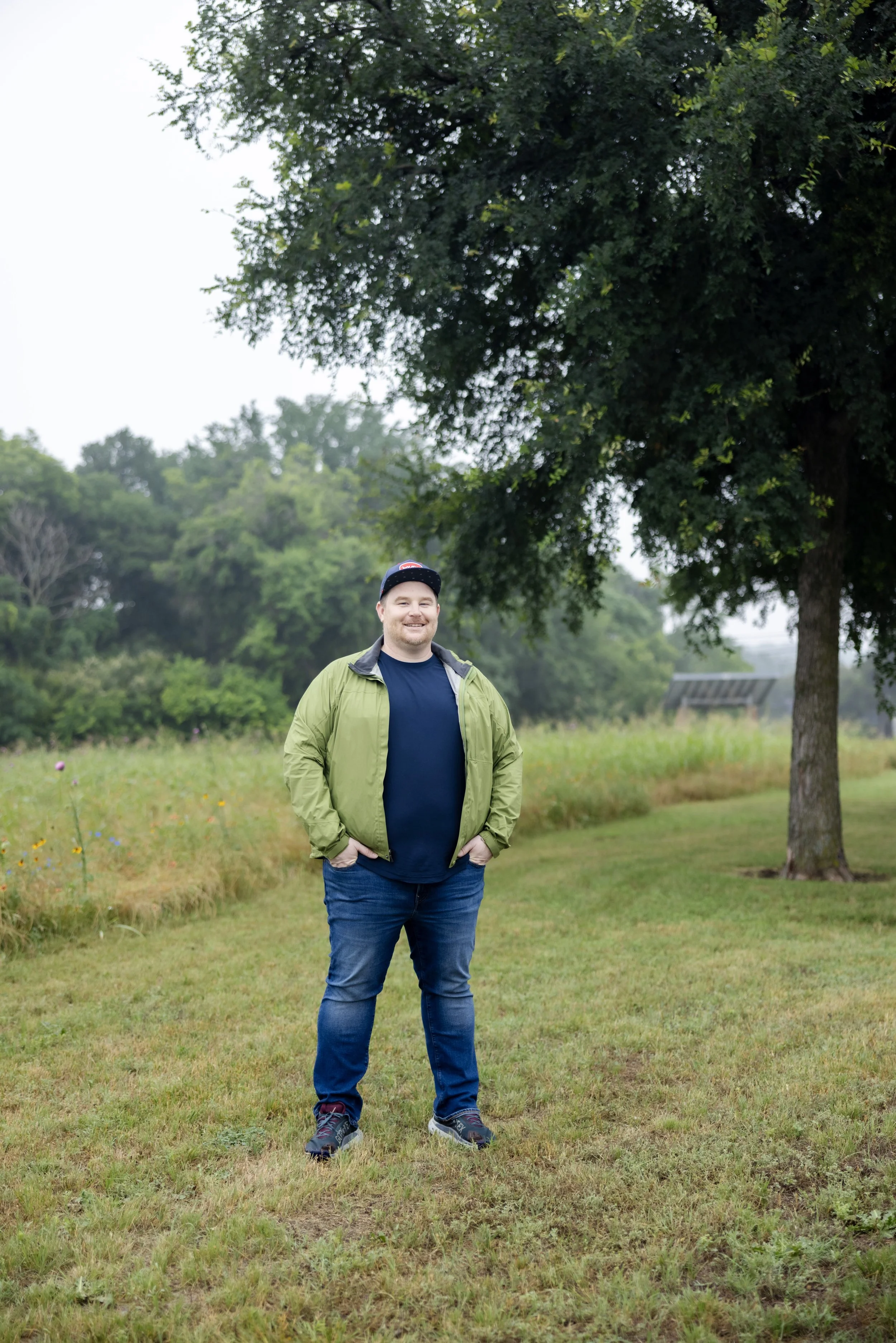 A man standing outdoors in a grassy area with trees and shrubs in the background, smiling and wearing a green jacket, navy shirt, jeans, and a cap.