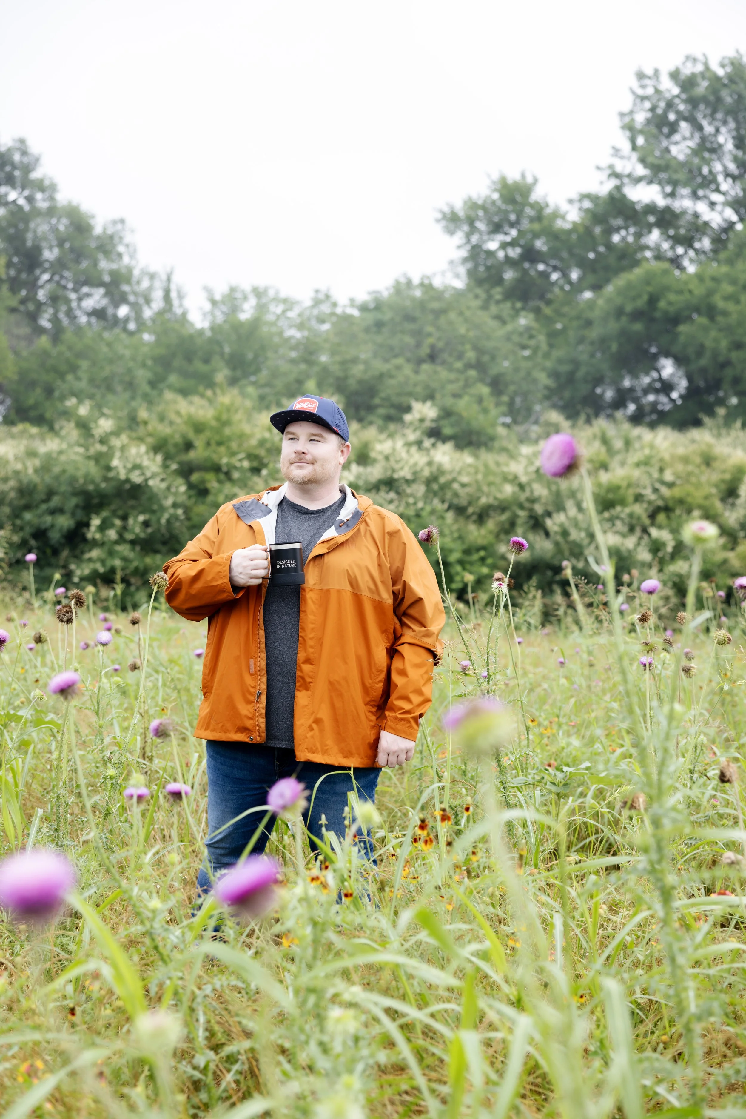 Man standing in a flower field holding a coffee mug, wearing an orange rain jacket, navy baseball cap, and gray t-shirt, surrounded by purple flowers and green trees in the background.