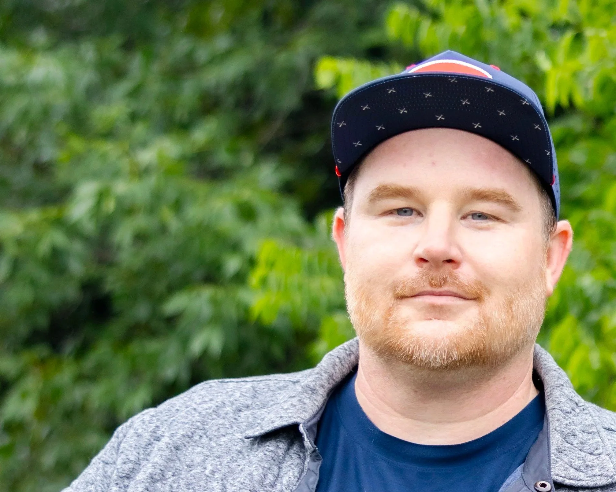 Young man with a red beard and blue eyes wearing a navy baseball cap and a gray hoodie outdoors with green foliage background.