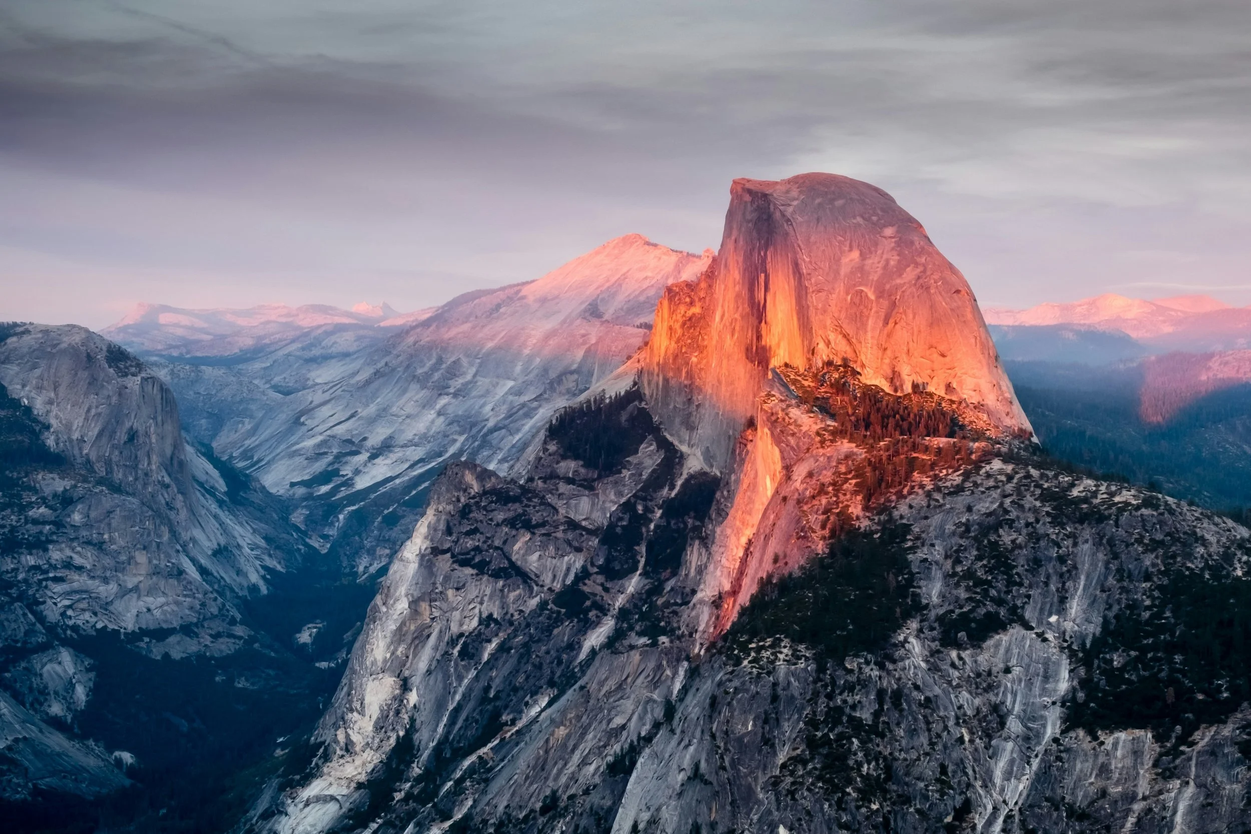 Sunset over a mountain range with a large granite formation, Half Dome, illuminated in orange light and surrounded by rugged peaks and valleys.