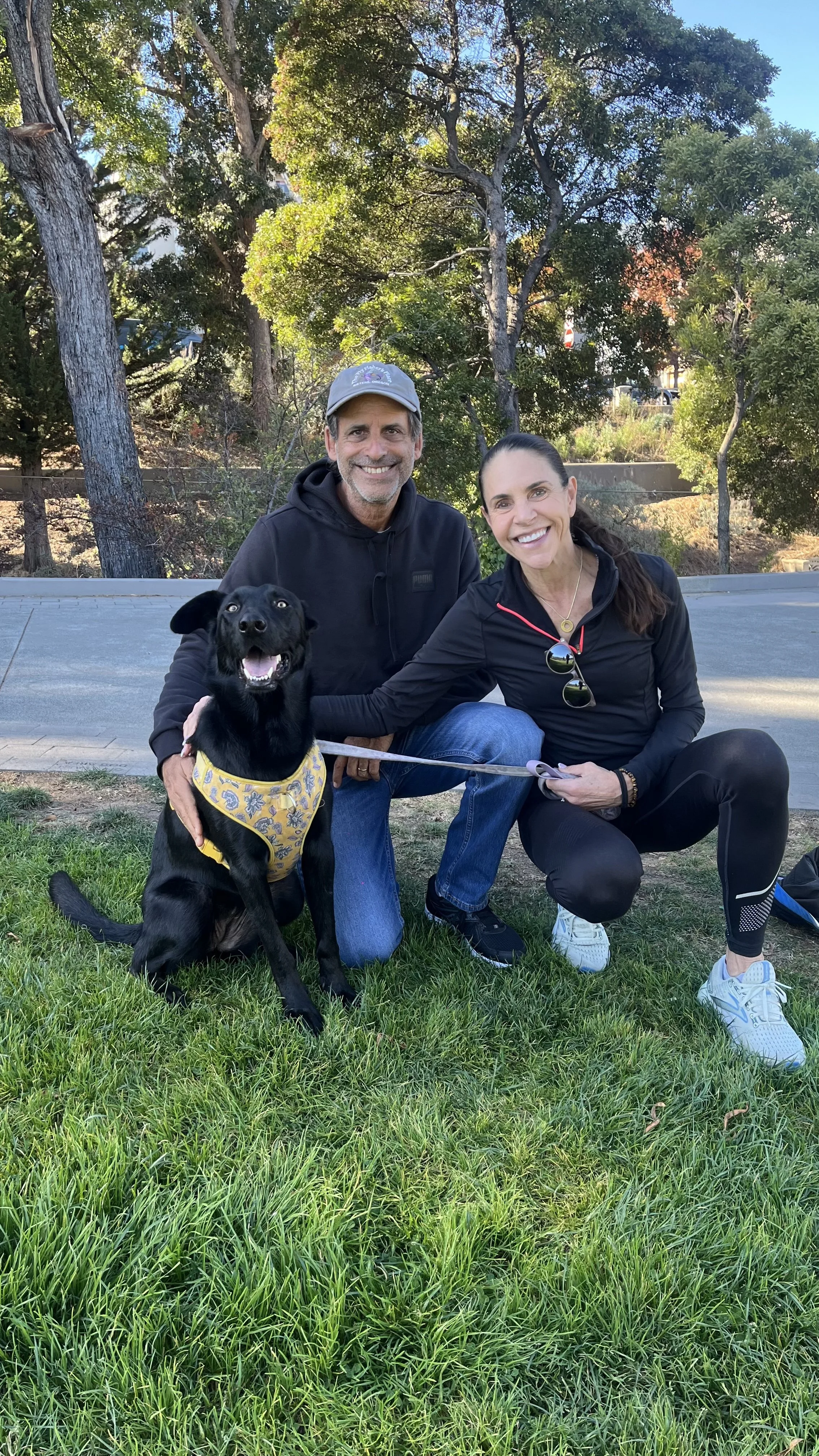Two smiling adults, a man and a woman, and a black dog with a yellow bandana posing outdoors on grass with trees in the background.