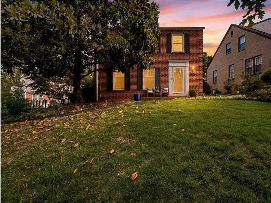 The image depicts a brick house with two stories, illuminated from inside. There are three chairs on the lawn near the house, and a large tree partially shading the yard. The sky in the background has a pinkish hue from sunset, and fallen leaves are scattered across the grassy yard.