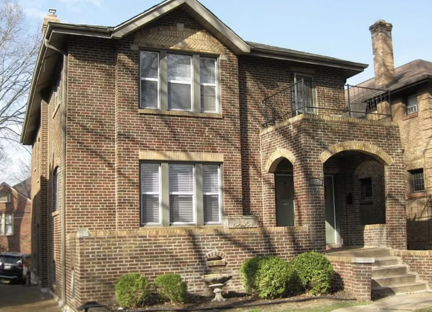 Brick three-story house with arched entrance, stairs, and windows on each level, surrounded by small bushes and a church fountain.