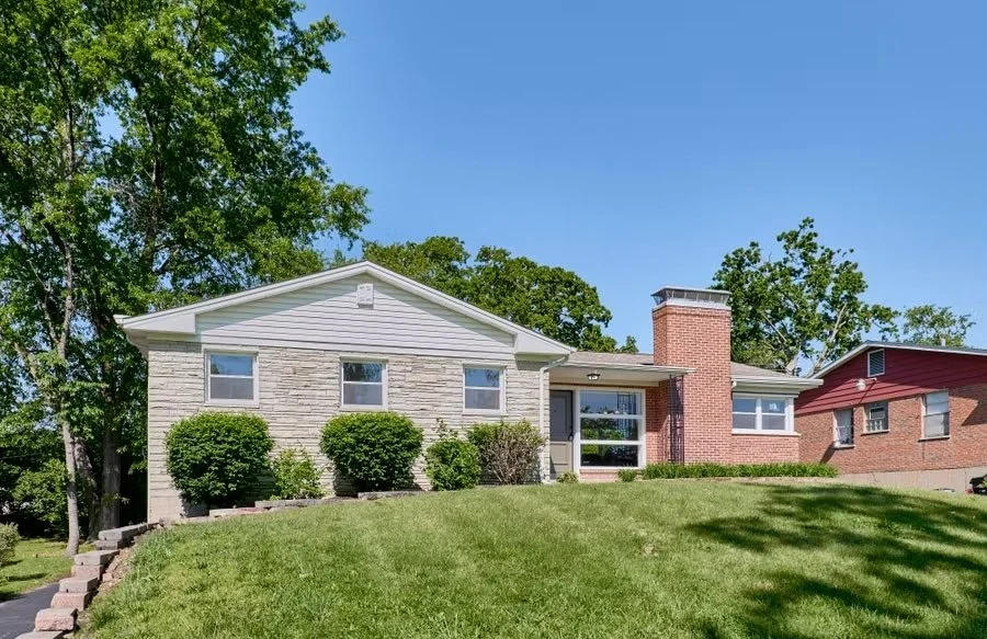 Front view of a house with a white brick exterior, large windows, a chimney, and a well-maintained lawn with bushes, under a clear blue sky.