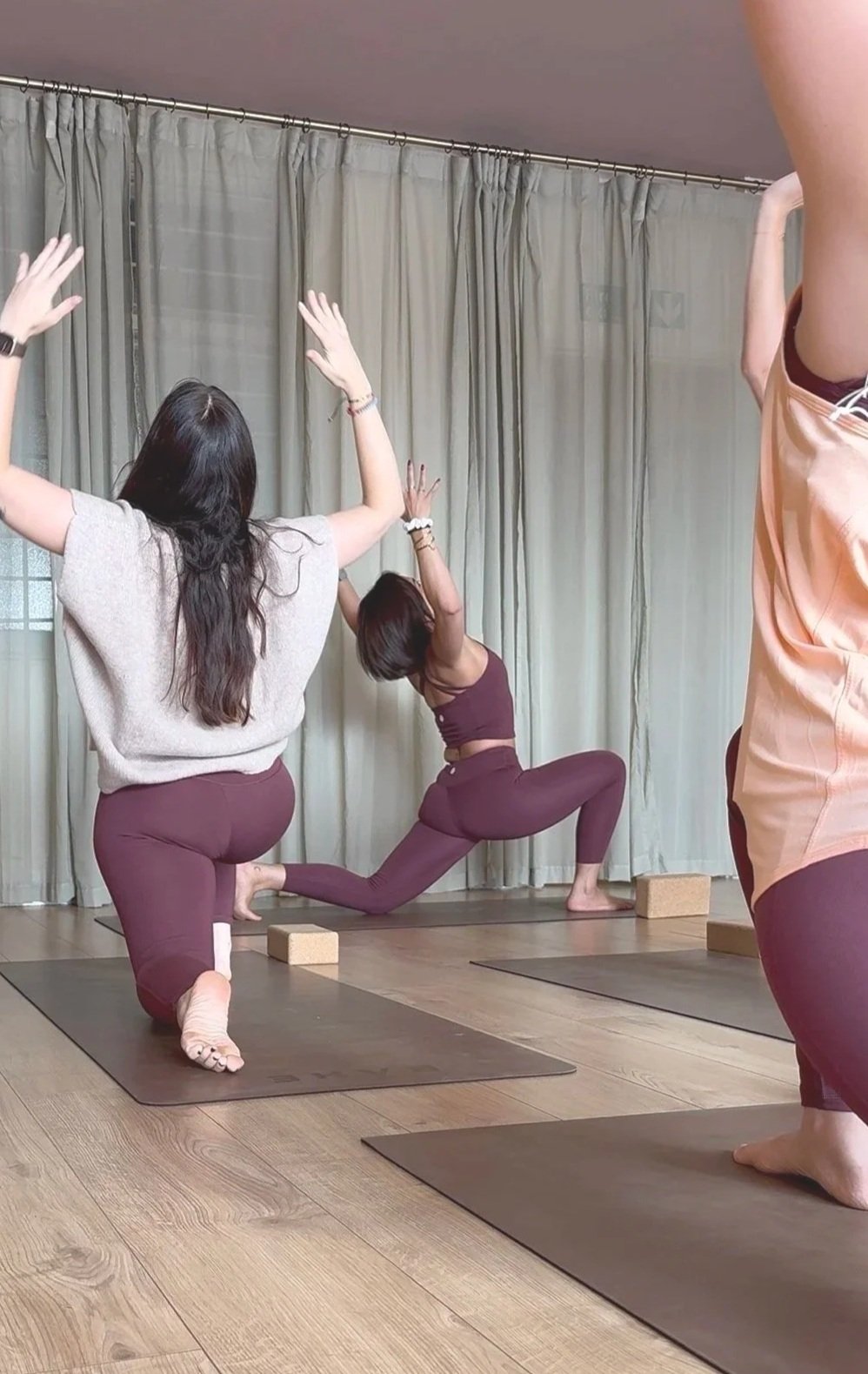 Three women practicing yoga indoors on mats with a curtain backdrop, one woman in a lunge pose with hands raised, two others kneeling or standing with arms raised.