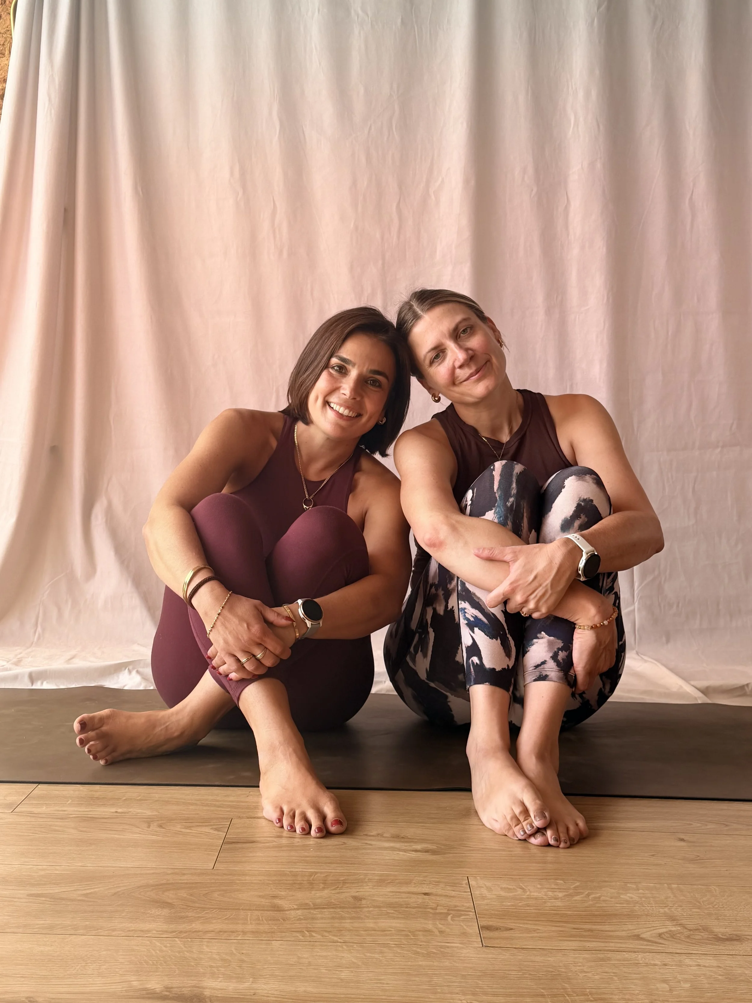 Two women sitting on a wooden floor with a beige backdrop, smiling, and hugging each other with their heads touching. They are barefoot with one woman wearing maroon and the other in patterned leggings.