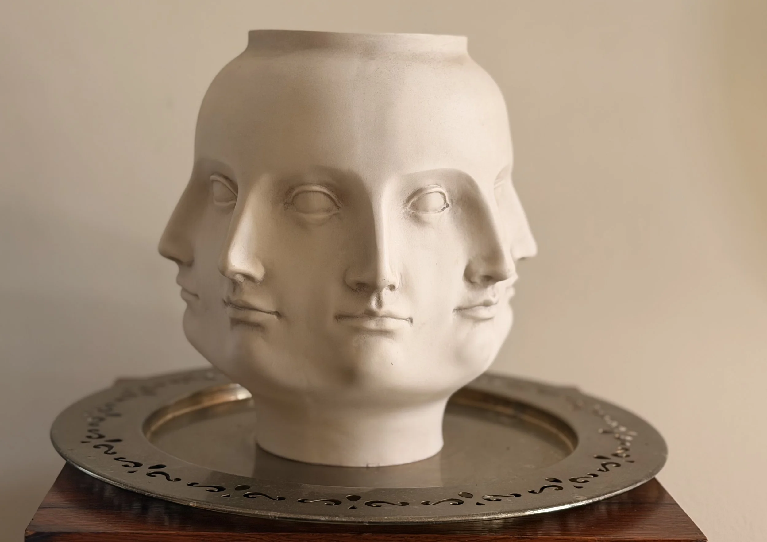 A decorative ceramic head sculpture resembling a human face, mounted on a circular metallic stand on a wooden surface.