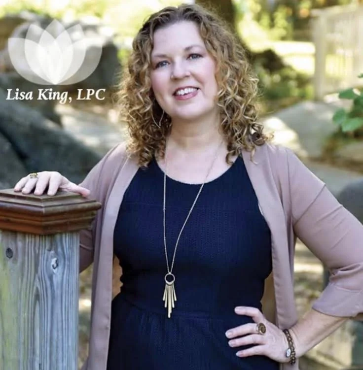 A woman with curly blonde hair wearing a navy dress, tan cardigan, and jewelry, standing outdoors with greenery, leaning on a wooden post, smiling at the camera.