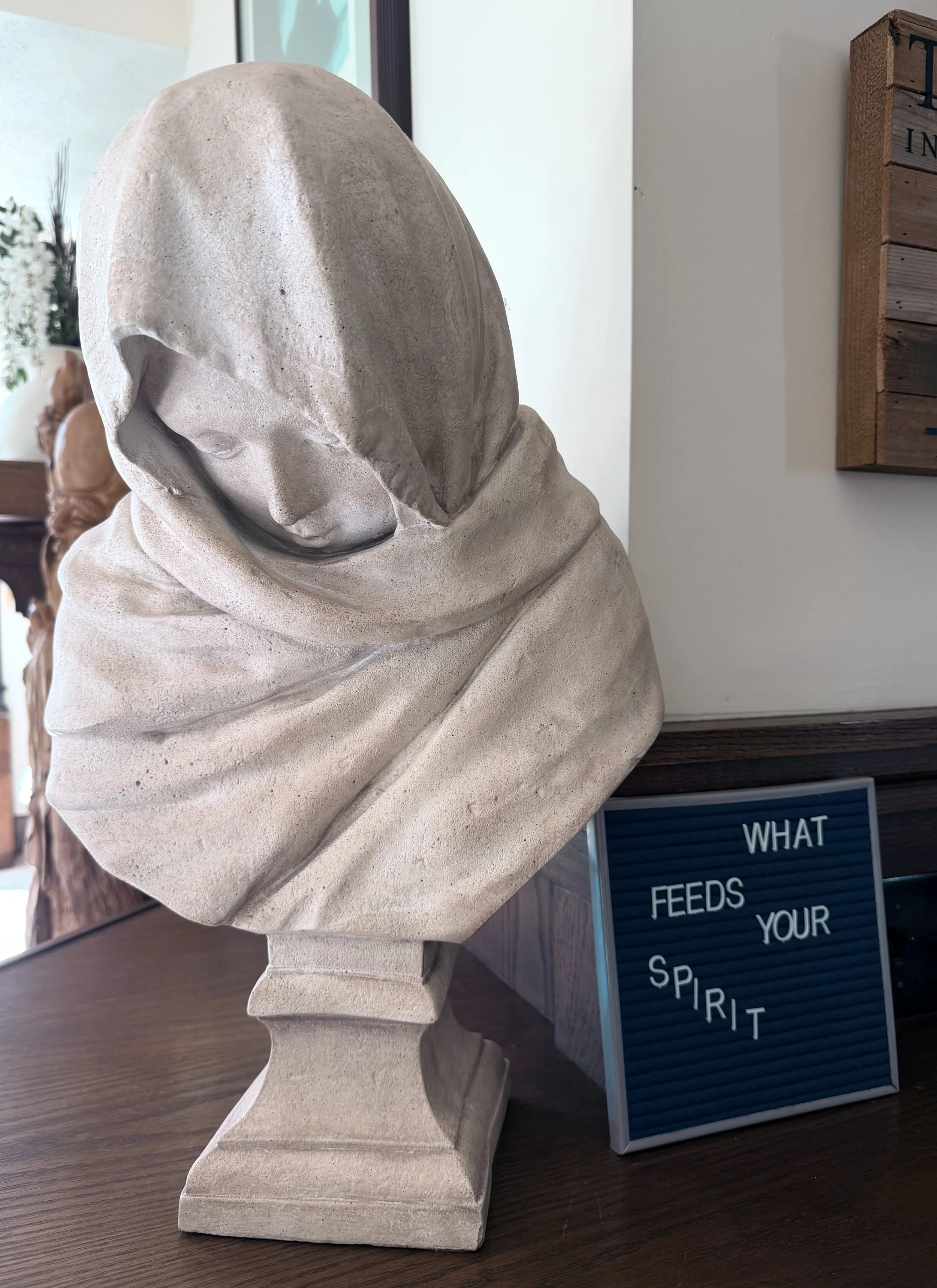 A white stone sculpture of a woman with a boulder-shaped head, with closed eyes and a peaceful expression, seated on a wooden surface. There is a sign with partial visible text in the background.