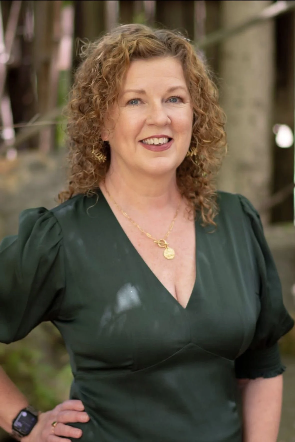 A woman with curly red hair, wearing a dark green dress and gold jewelry, standing outdoors with trees in the background.