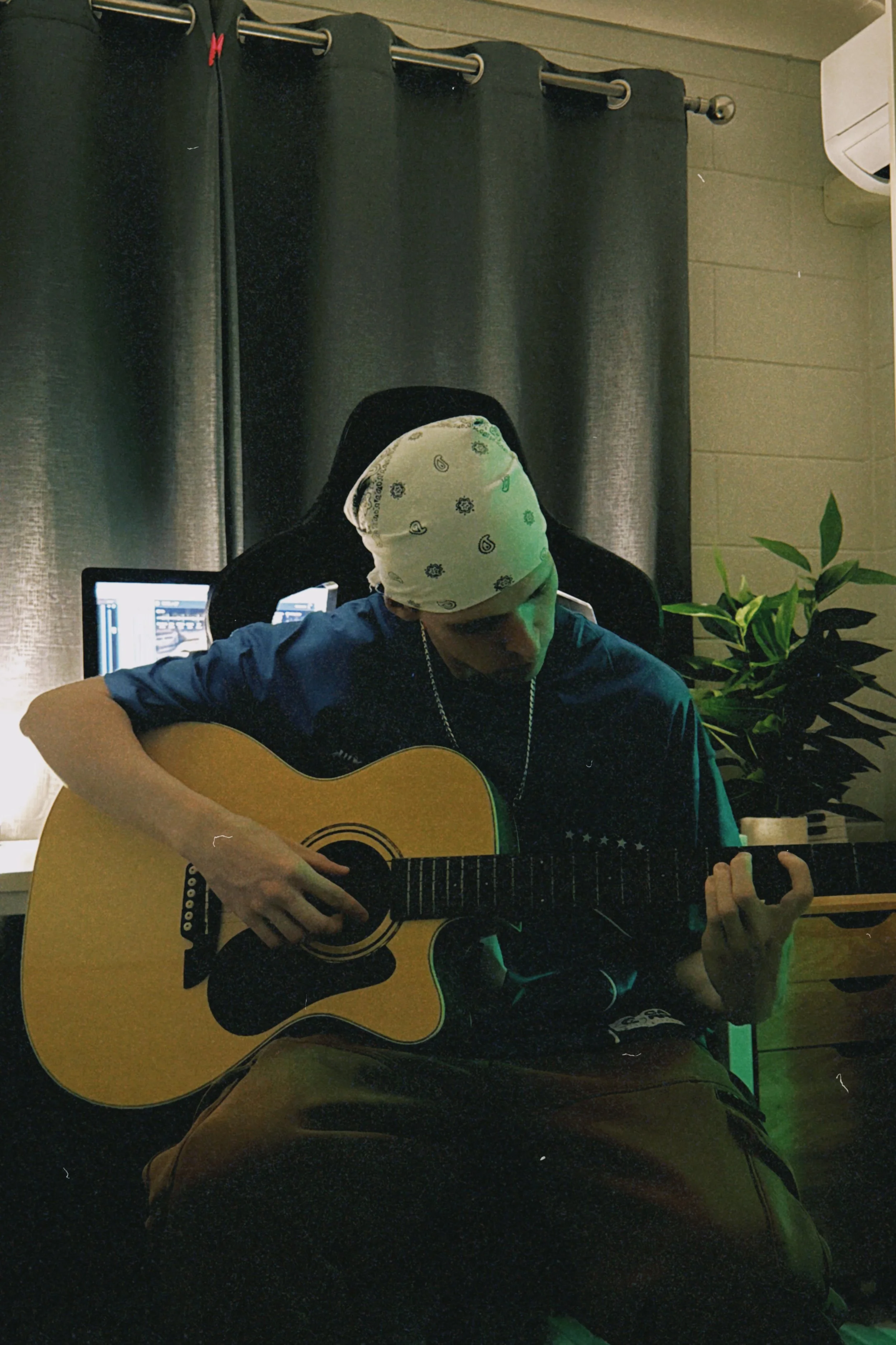A person with a white bandana on their head, sitting indoors and playing an acoustic guitar. Black curtains, a computer screen, and a potted plant are visible in the background.