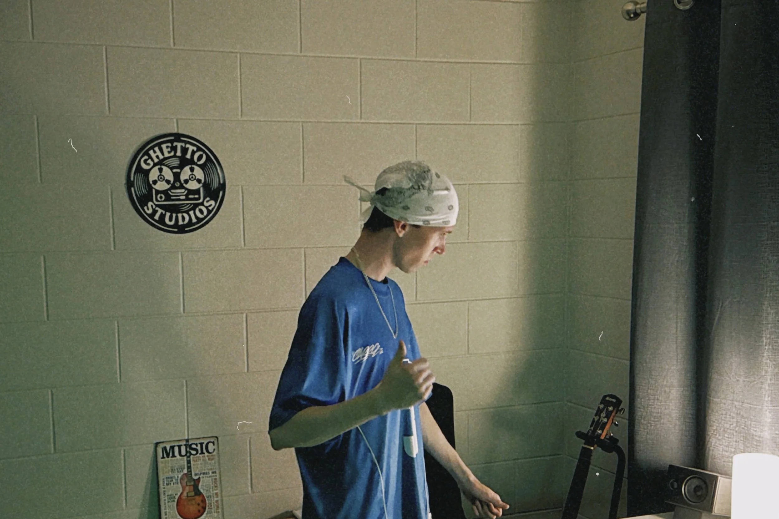 A young man wearing a bandana and a blue shirt with white writing, listening to white earphones and holding a drink, standing next to a guitar on a stand against a beige brick wall with musical posters.