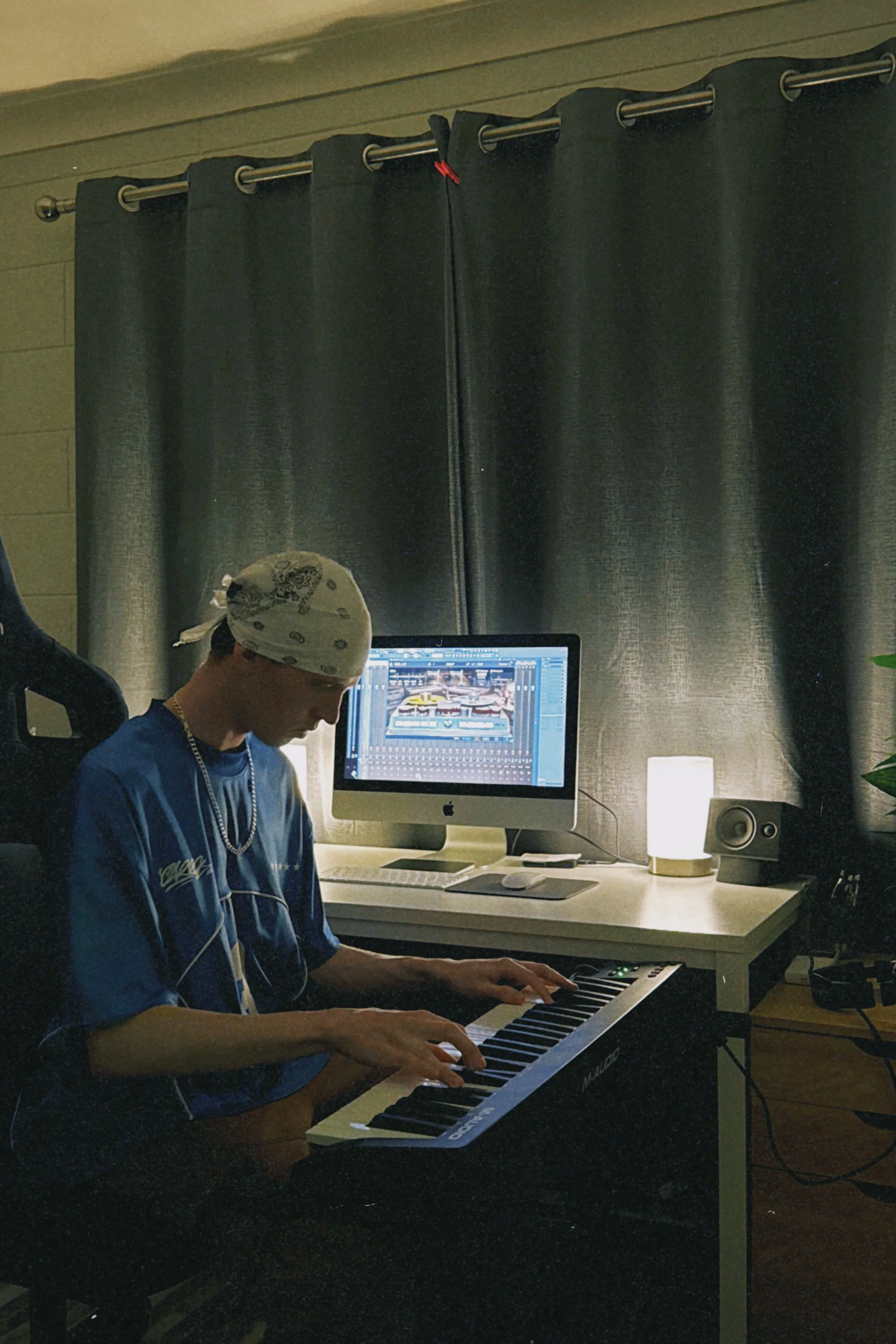 Person wearing a bandana playing a digital keyboard in a home studio with a computer monitor and speakers, dark curtains in the background.