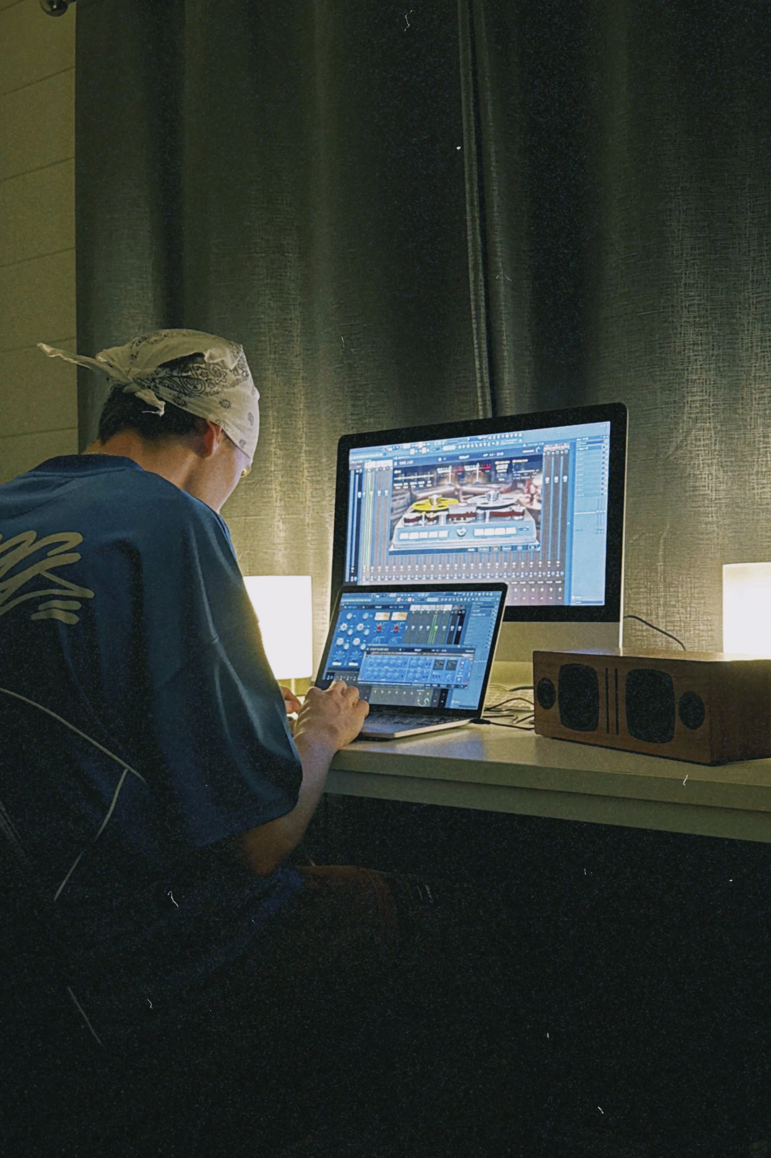 Person working on music production with multiple computer screens, one displaying a DJ software interface, in a dimly lit room.