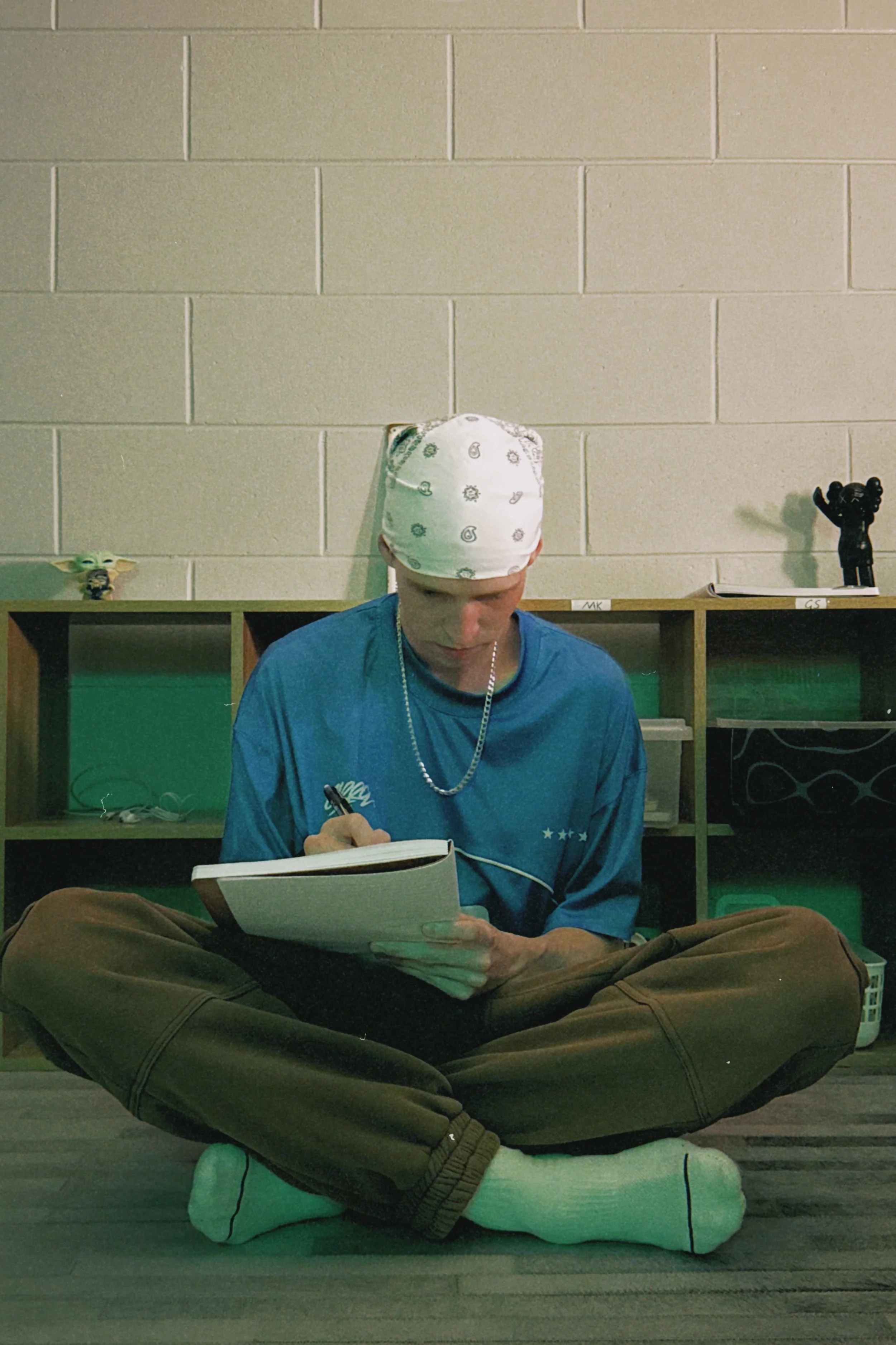 Person sitting cross-legged on the floor, reading a notebook, wearing a white bandana with a paisley pattern, a blue shirt, and brown pants, with a green-background shelf and yellow wall behind.