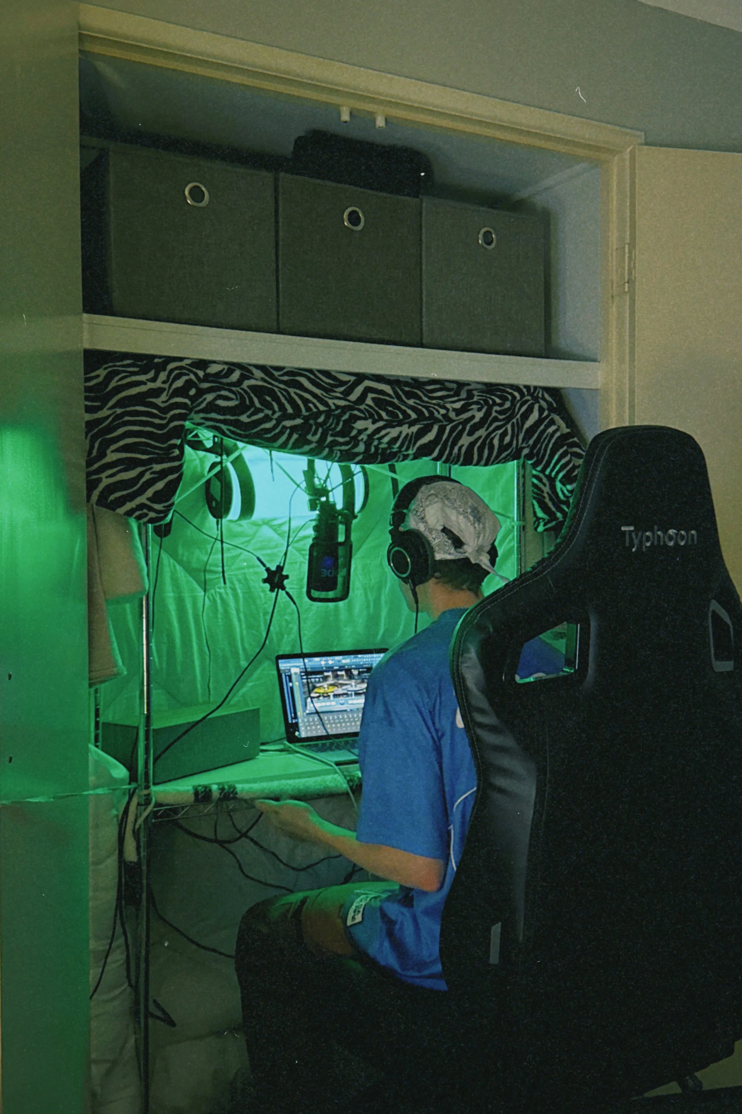 A person wearing headphones and a blue shirt is sitting at a desk with a laptop, inside a green screen studio setup with a zebra-patterned fabric and shelves above with storage boxes.
