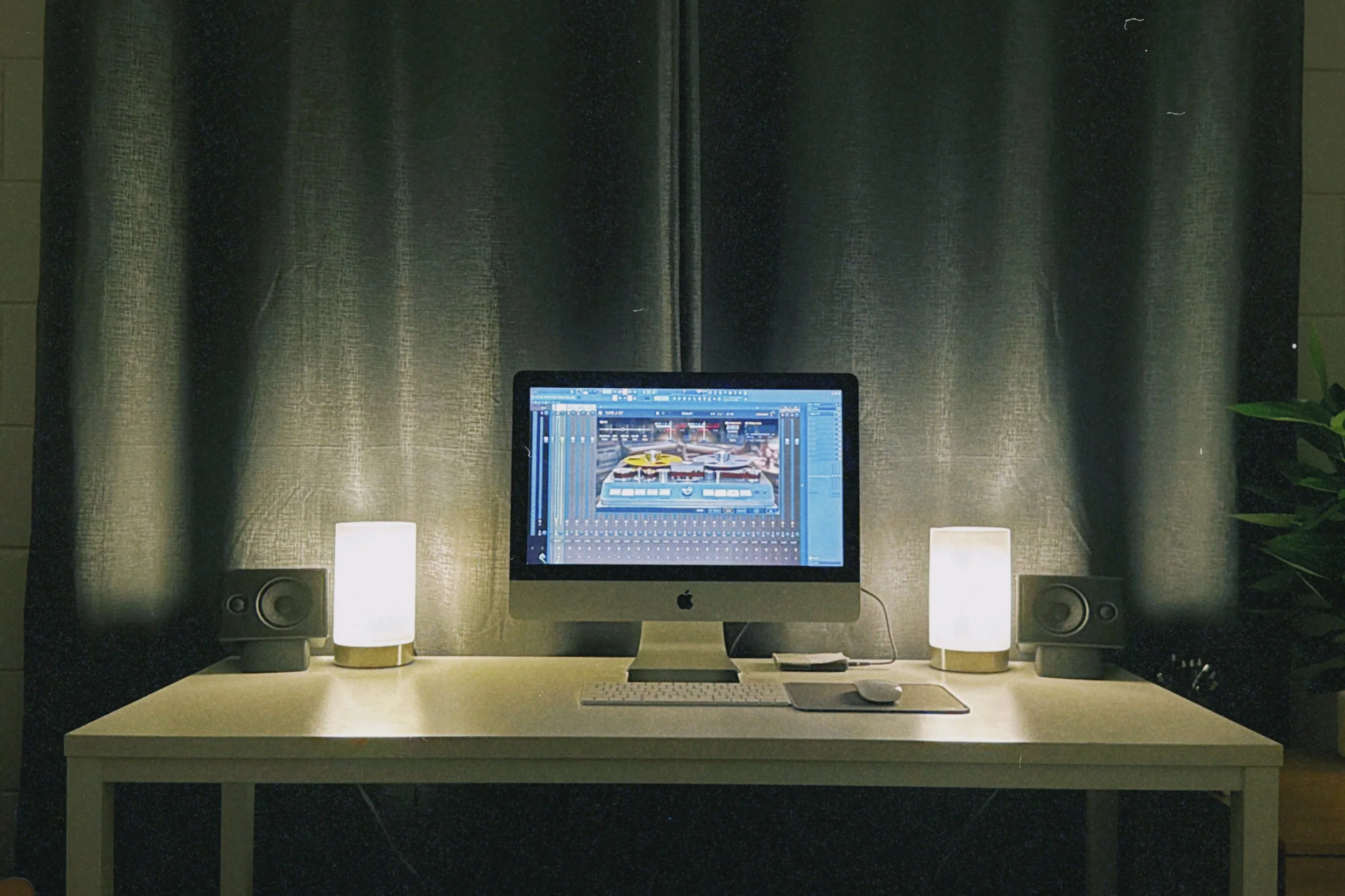 A computer desk setup with an iMac displaying a digital audio workstation, flanked by two white speakers with white bases, illuminated by two small table lamps, with a green plant on the right and a black curtain in the background.