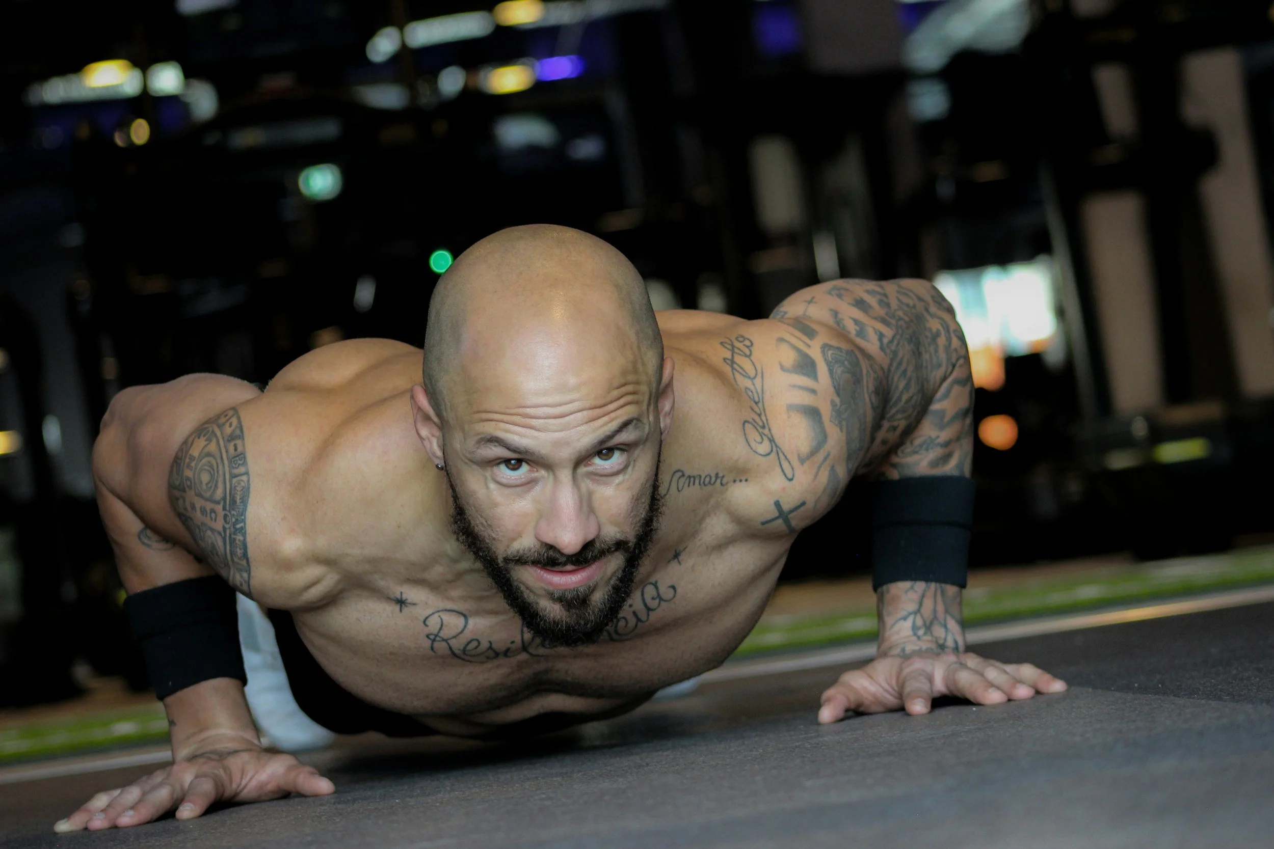 A muscular, bald man with tattoos performs a push-up in a gym, looking focused and determined.