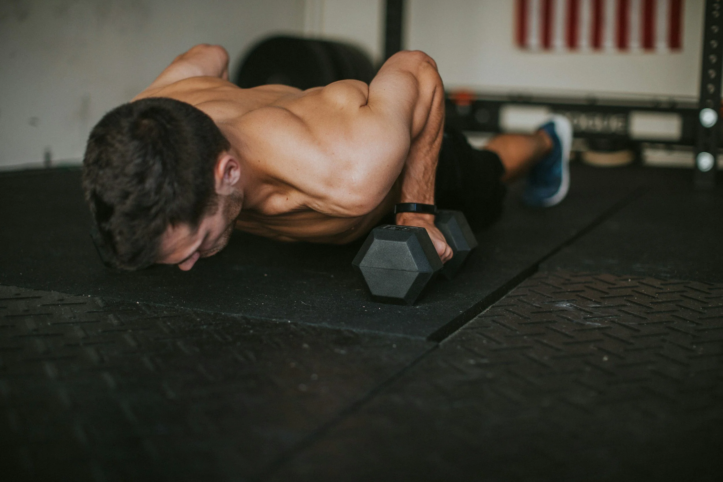A shirtless man doing a plank exercise on a black rubber gym floor, supporting his upper body with both arms resting on dumbbells, with an American flag in the background.