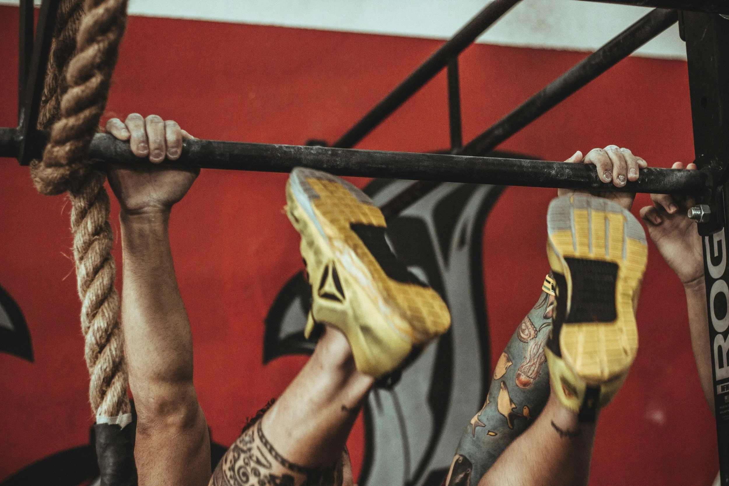 People hanging upside down from a pull-up bar, wearing colorful athletic shoes and shorts, with a red wall in the background.