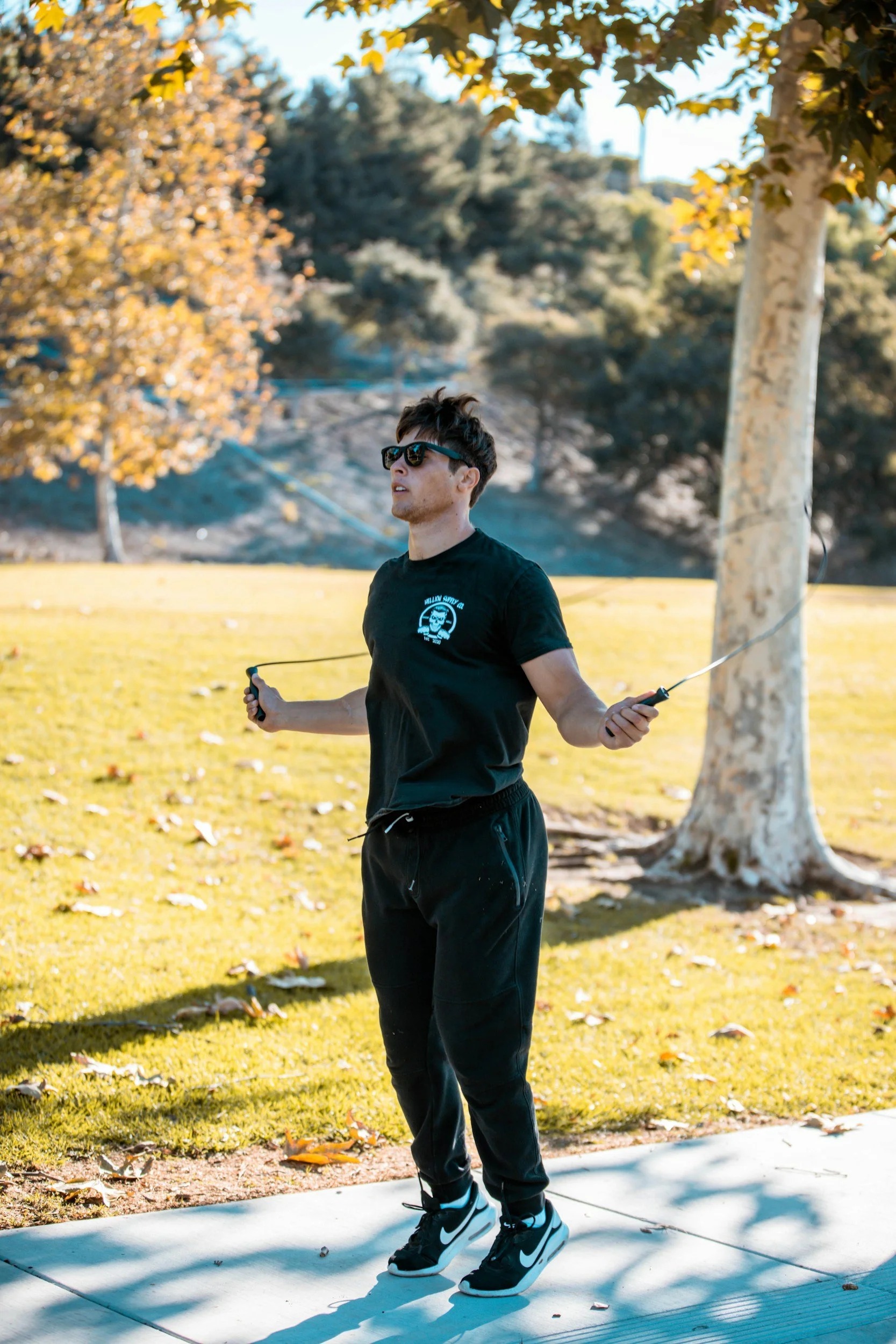 A young man wearing sunglasses, a black T-shirt, and black pants skipping with a jump rope outdoors in a park during fall.