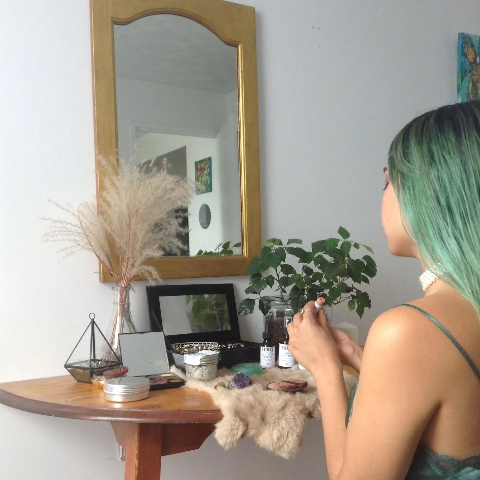 A woman with green hair, wearing a pearl choker and a teal camisole, sitting at a wooden vanity with various jewelry and skincare items, looking into a mirror on the wall.