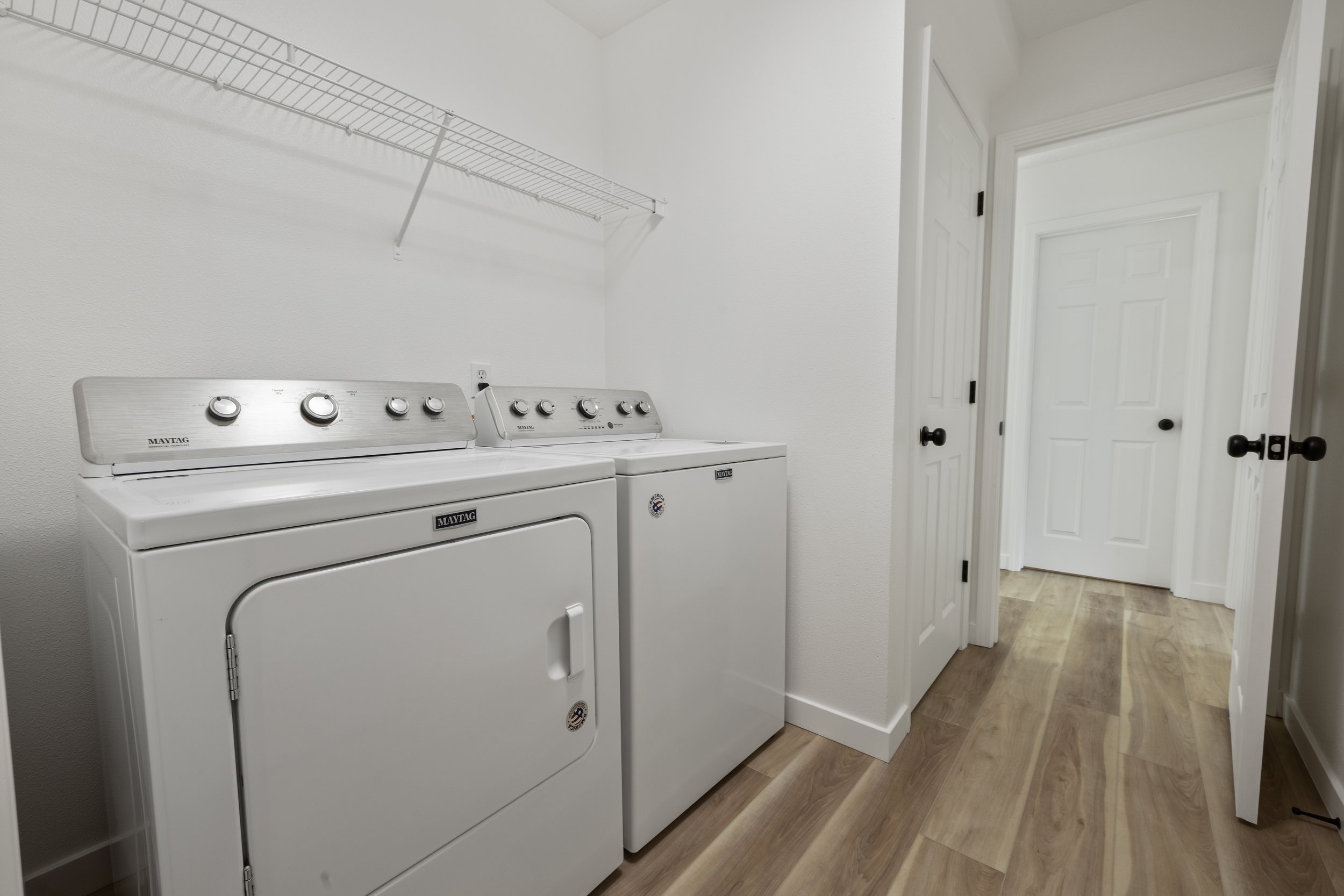 A laundry room with a white washing machine and dryer, a wire shelf above them, white walls, wood flooring, and a door leading to another room.