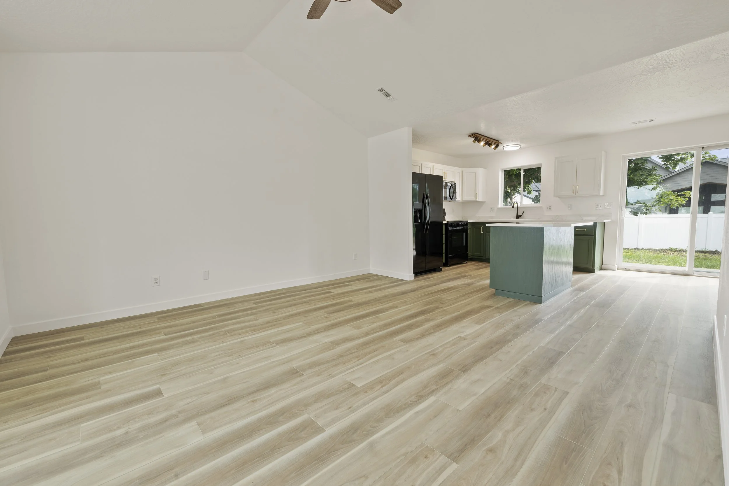 Bright open-concept living space with light-colored wooden flooring, white walls, and a modern kitchen with white cabinets, black appliances, and a kitchen island with a green base. Large windows and sliding glass door bring in natural light and offe