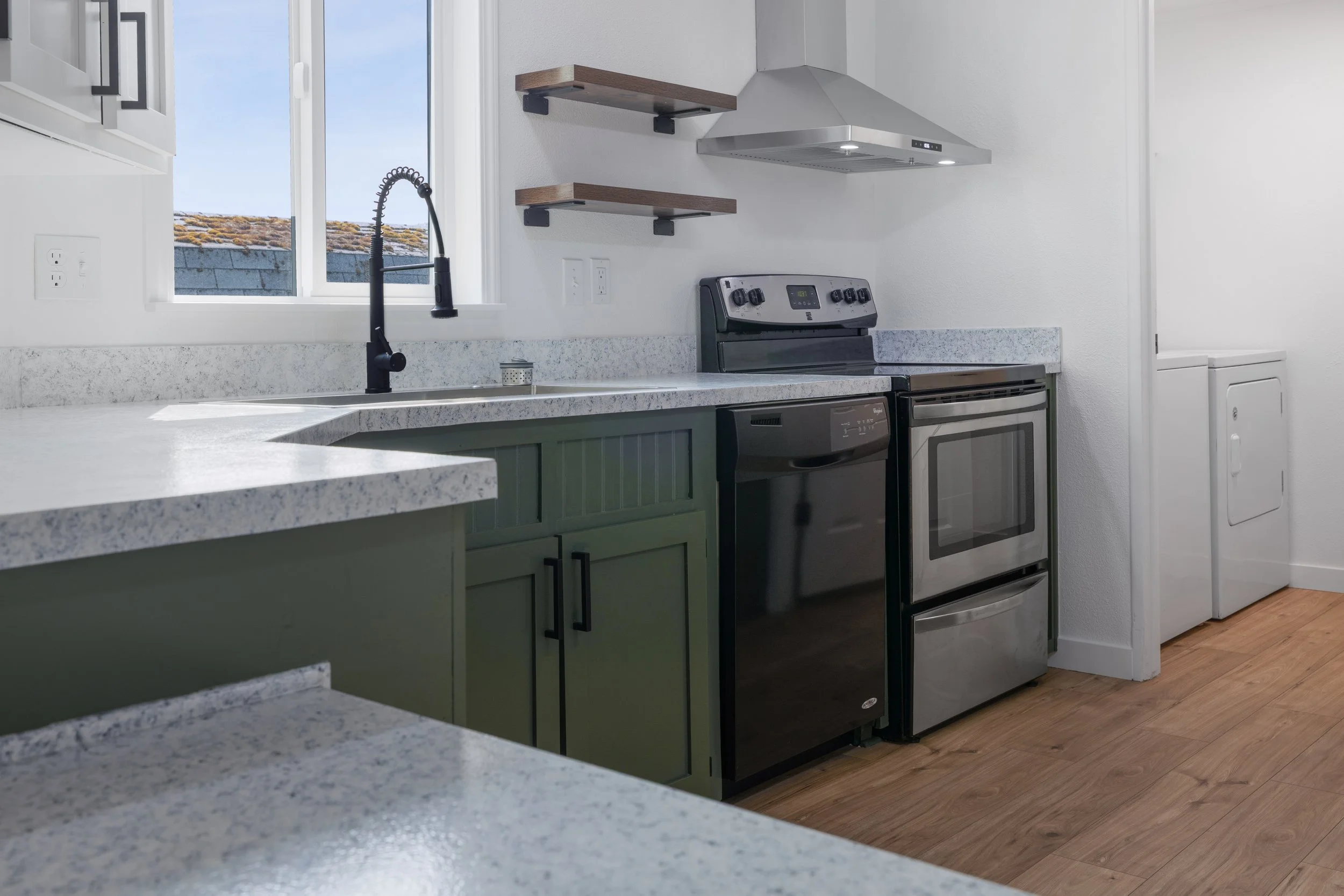 Modern kitchen with granite countertops, black faucet, stainless steel stove, white walls, open window showing a blue sky and roof, and hardwood floors.