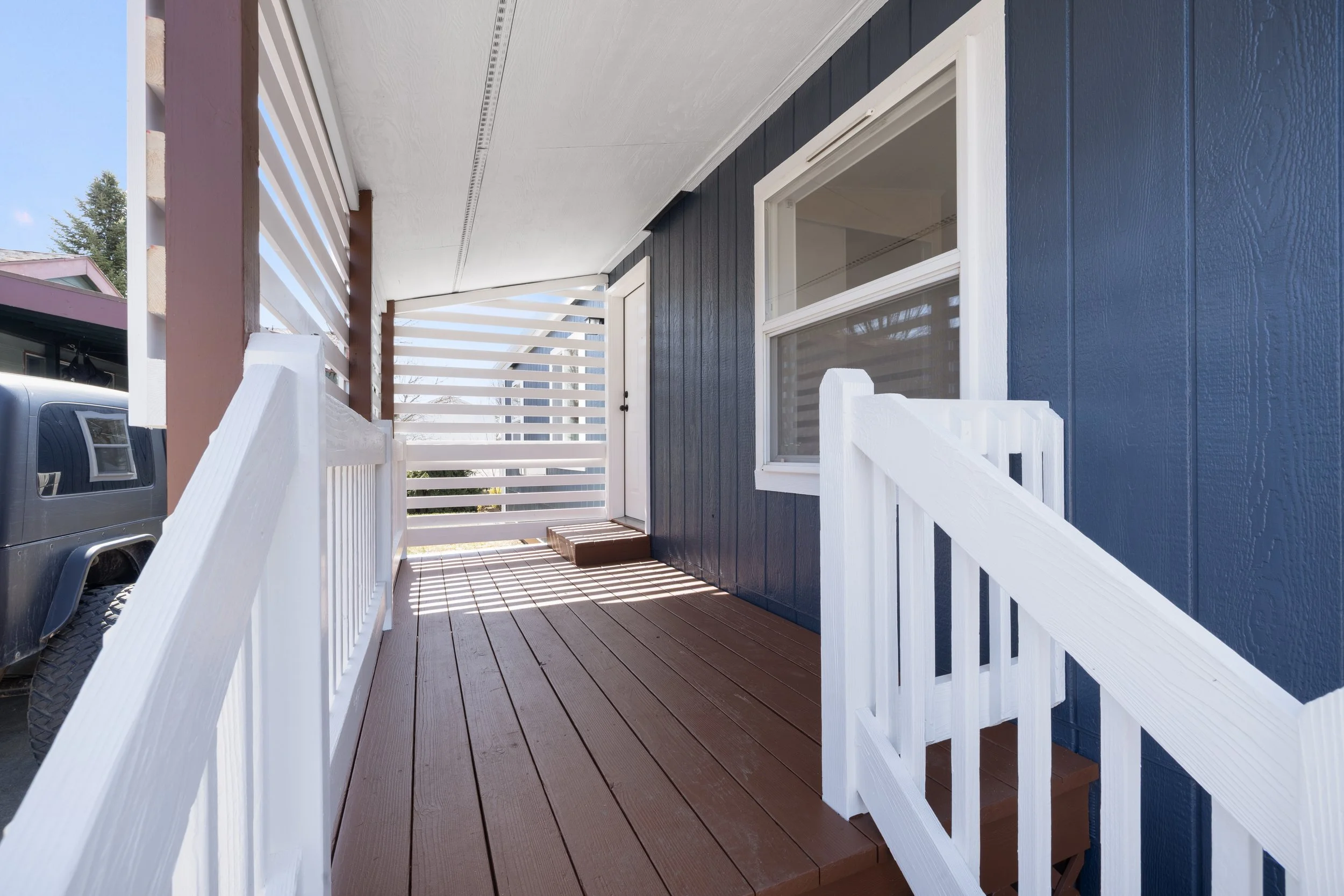 Front porch with brown wooden flooring, white railing, blue house wall, and white window, with a screen door and stair steps.