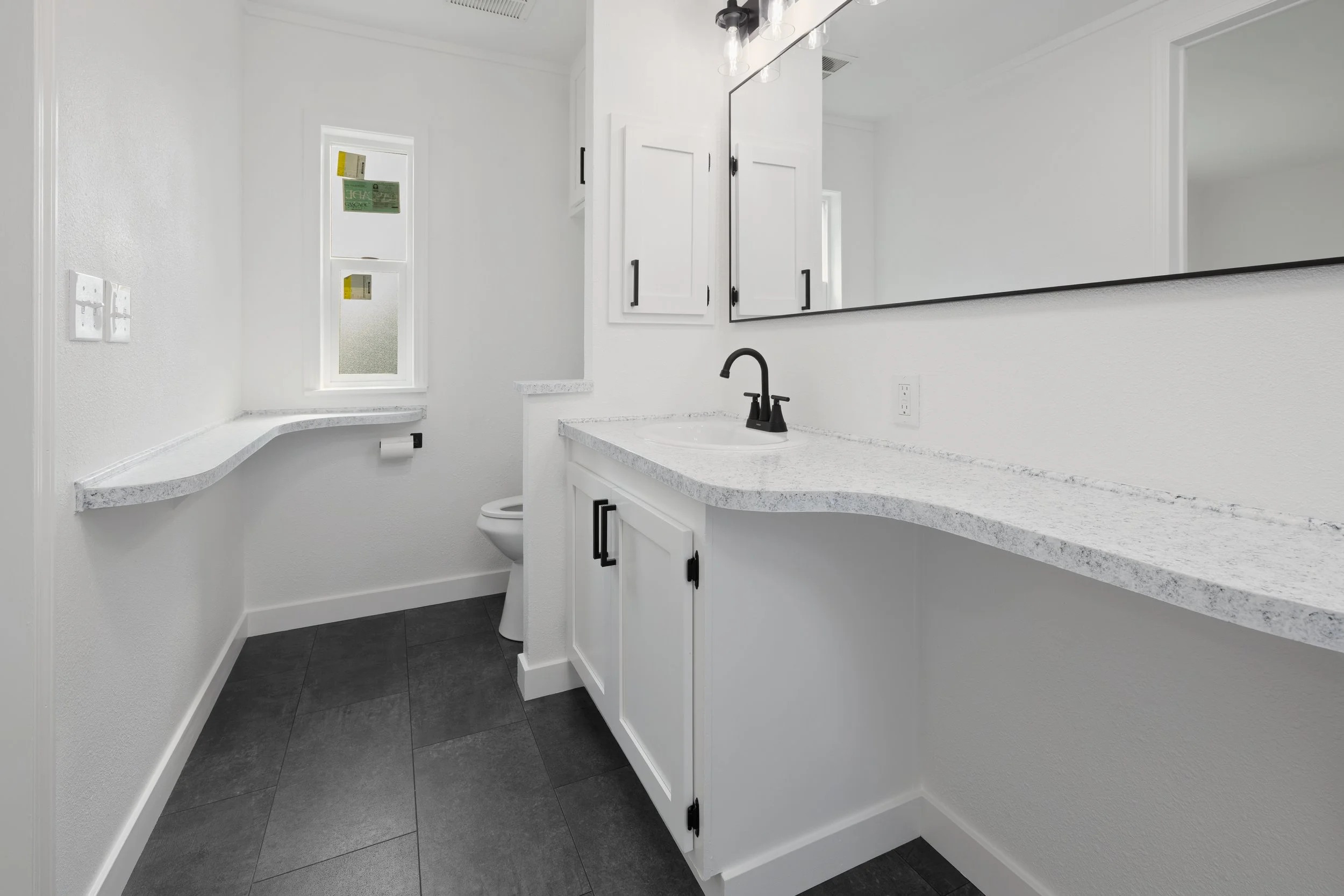 A modern, minimalistic bathroom with white walls, dark floor tiles, a large mirror, a white cabinet with black handles, a black faucet, and a white countertop with granite pattern.