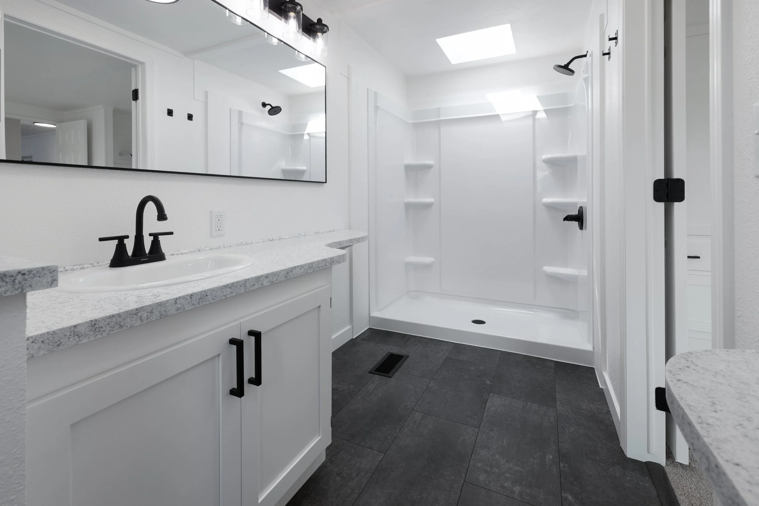 A modern bathroom with white walls, a gray speckled countertop with a white sink and black fixtures, a large mirror, black cabinet handles, a shower with black fixtures and shelves, and a skylight letting in natural light.