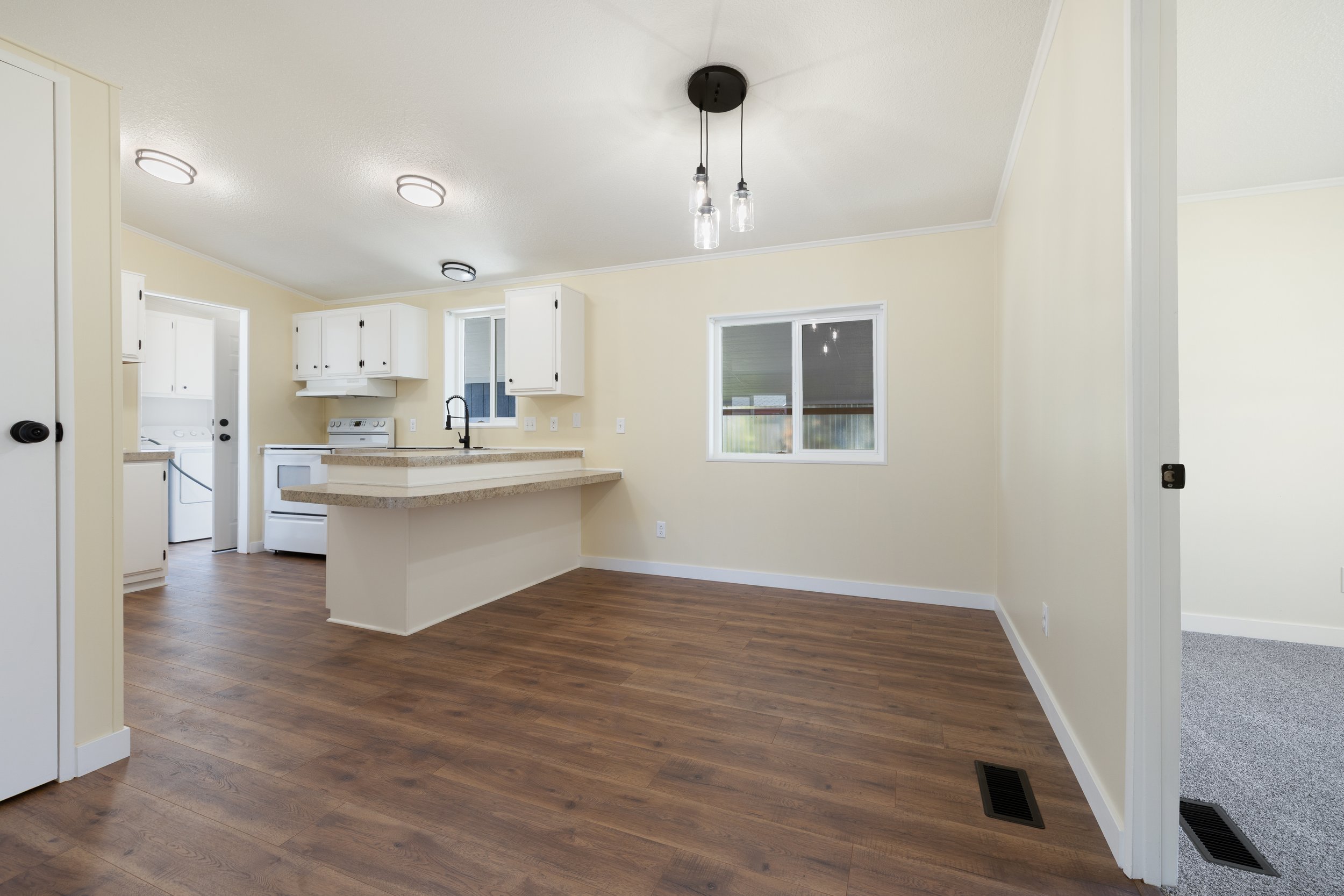 Empty kitchen and living space with wood flooring, white cabinets, and a large window.