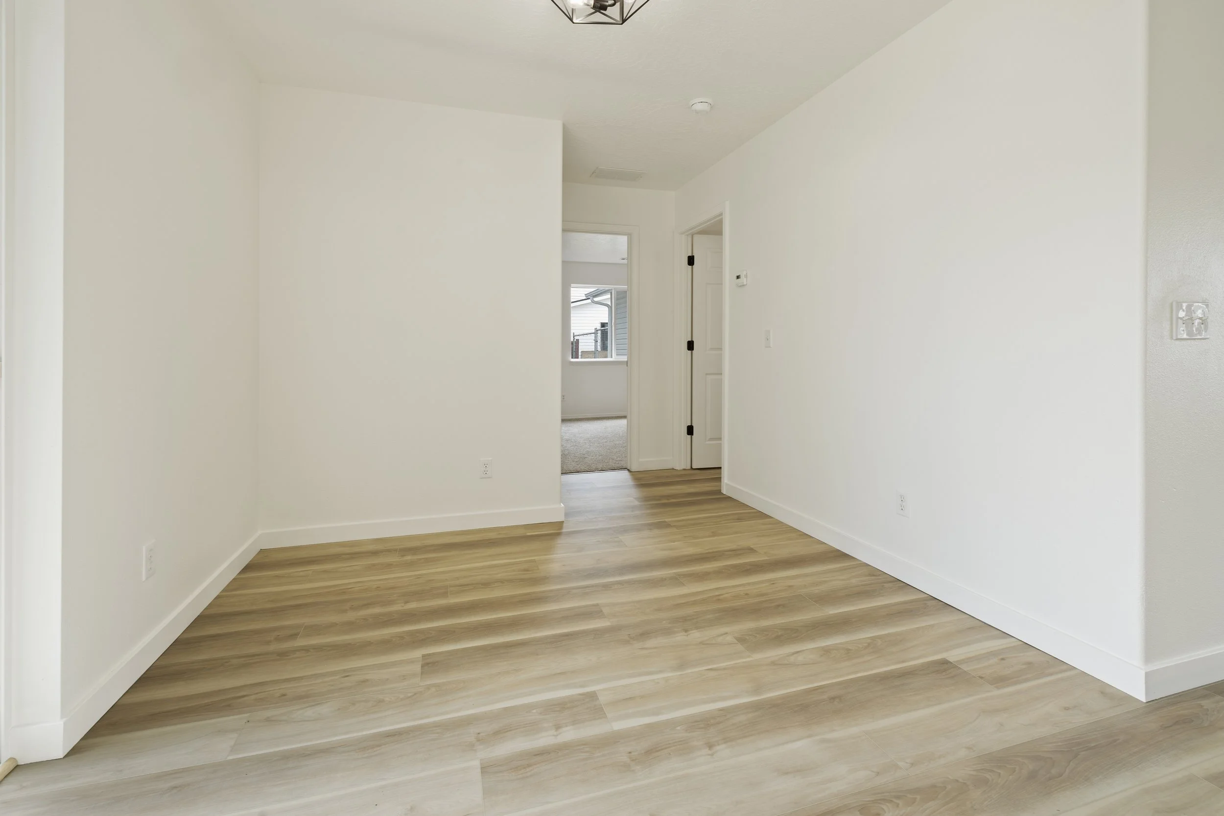 Empty room with white walls and wooden flooring, showing a doorway leading to another room with a window.