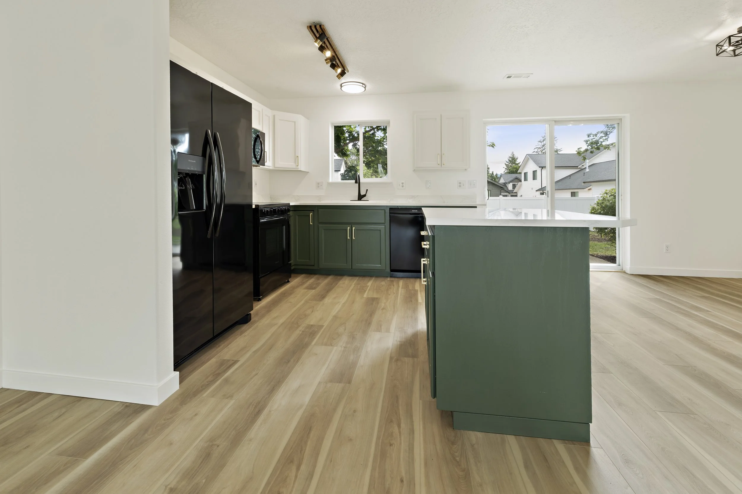 Modern kitchen with white upper cabinets, dark green lower cabinets, black refrigerator and appliances, and large windows overlooking neighboring houses and trees.