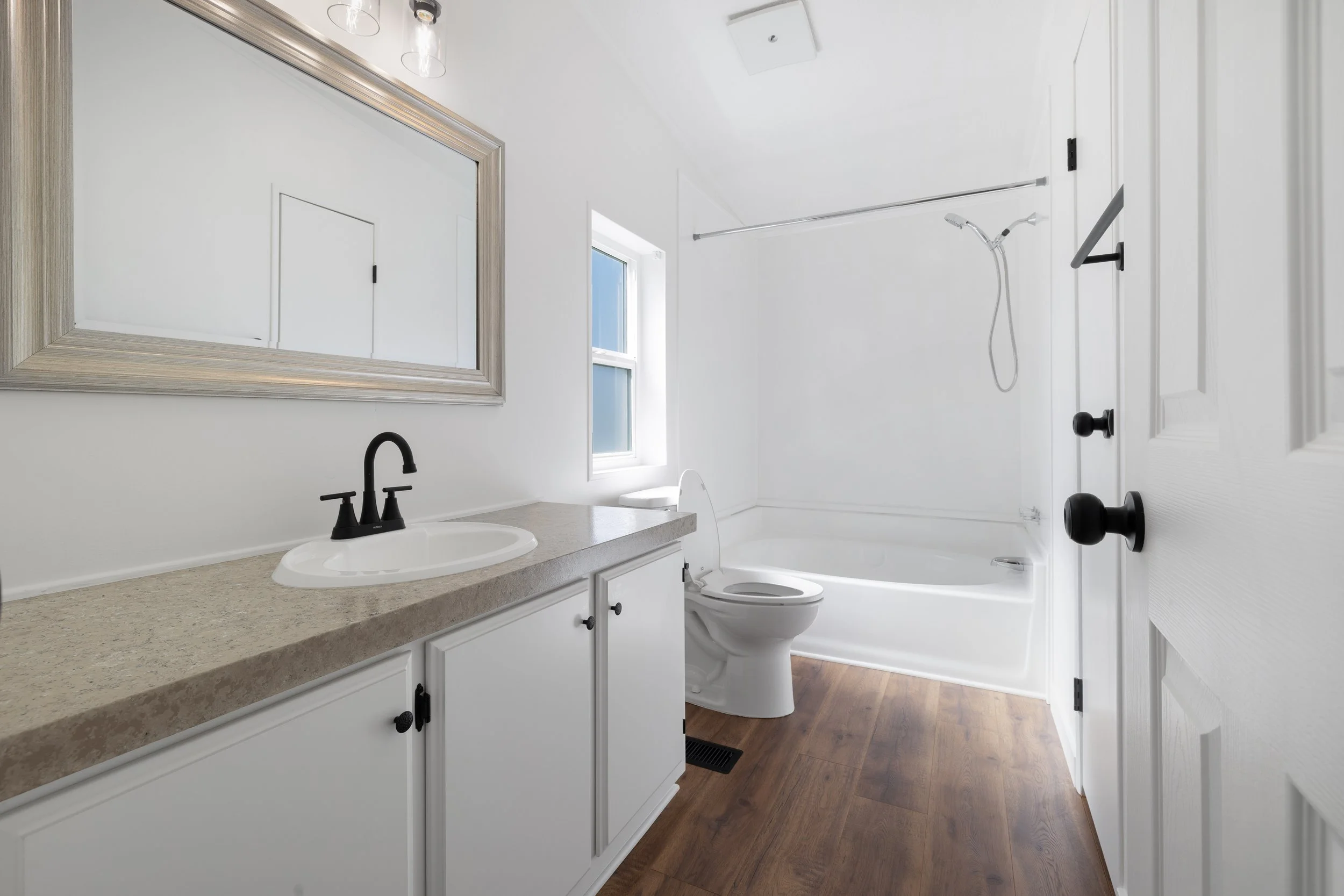 A bathroom with a white vanity, beige countertop, black faucet, large mirror, and a shower area with a white bathtub, showerhead, and glass window. The room has white walls, a wooden floor, and a door with black handle.
