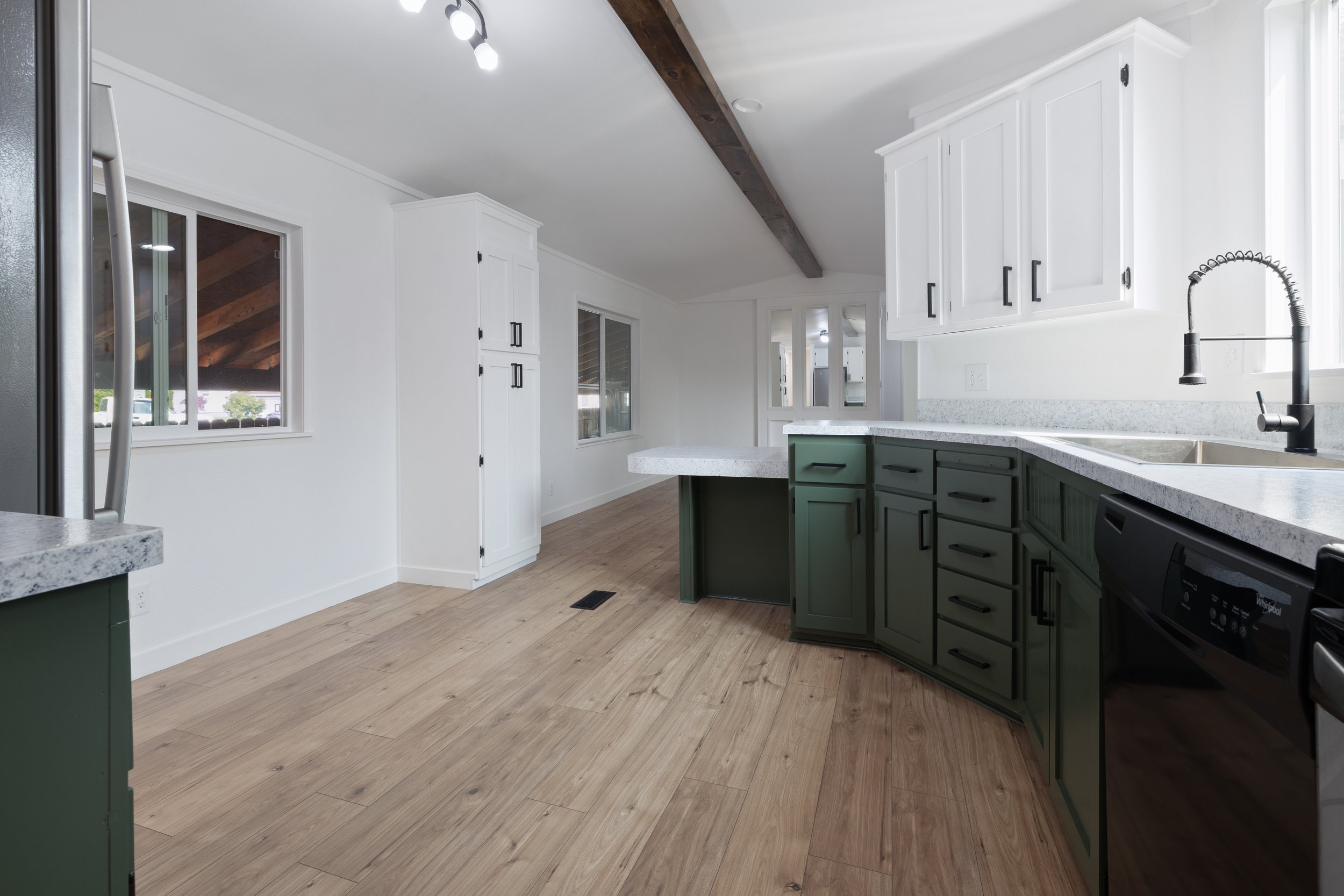 Modern kitchen with white upper cabinets, olive green lower cabinets, white speckled countertops, stainless steel faucet, light wood flooring, and large window letting in natural light.