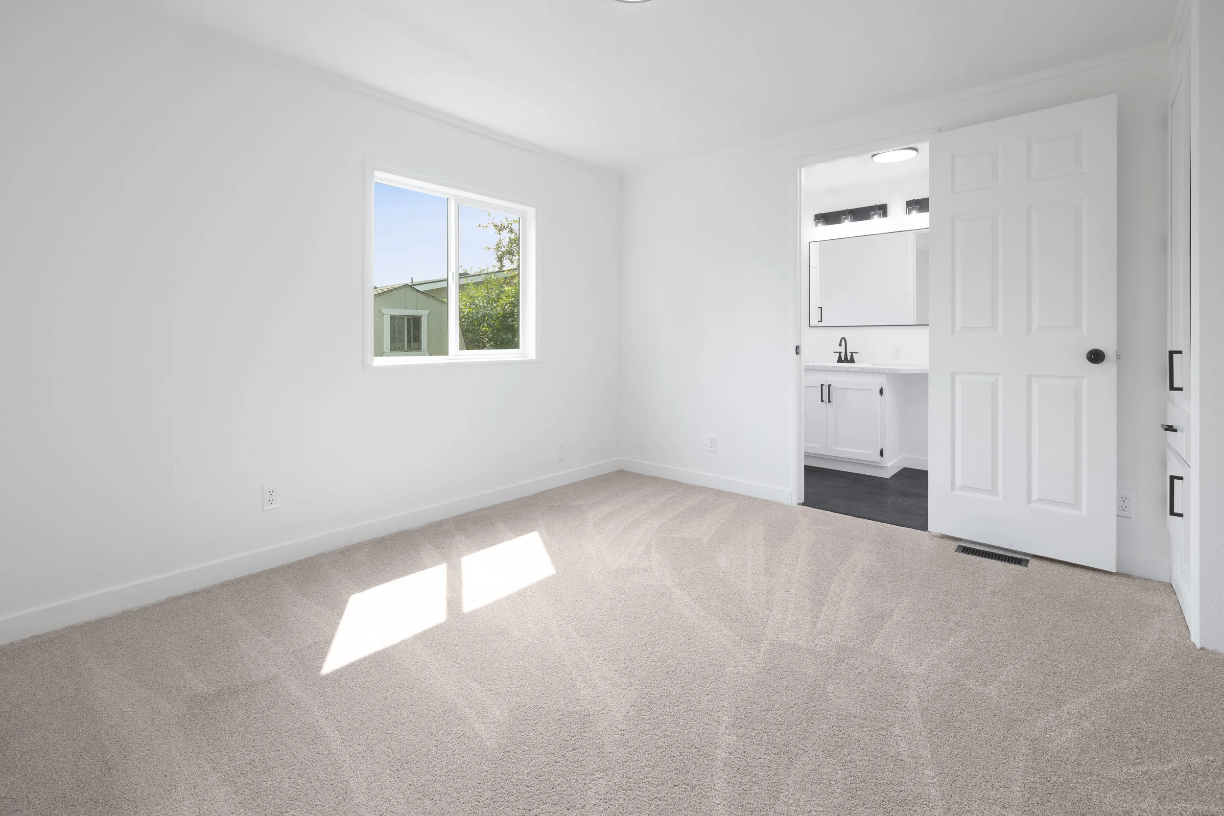 Empty room with beige carpet, white walls, a window showing trees outside, and a doorway leading to a bathroom with a mirror and dark tile floor.