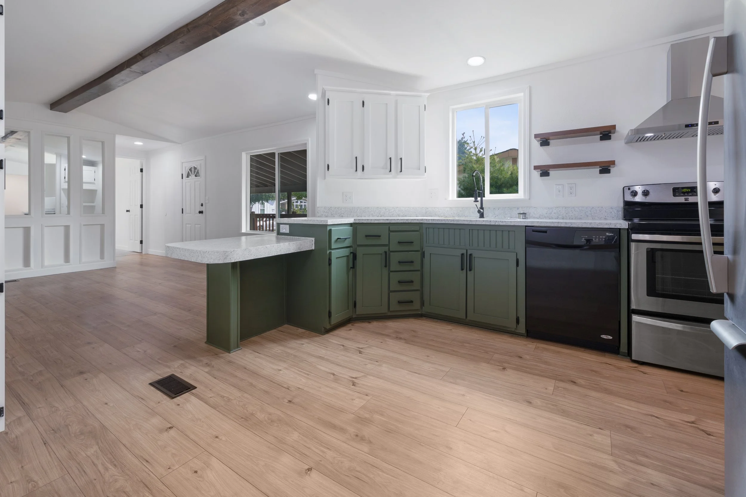 Bright kitchen with white walls, green cabinets, and light wood flooring. It features a window, stainless steel appliances, open shelves, and a countertop with a sink.