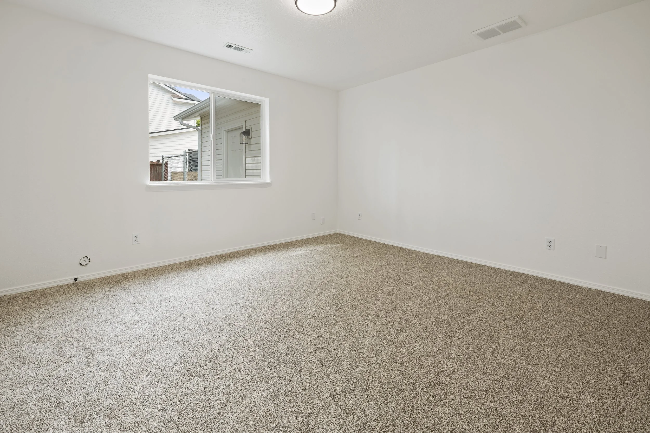 Empty white room with a window showing outdoor view, beige carpet, and ceiling lights.